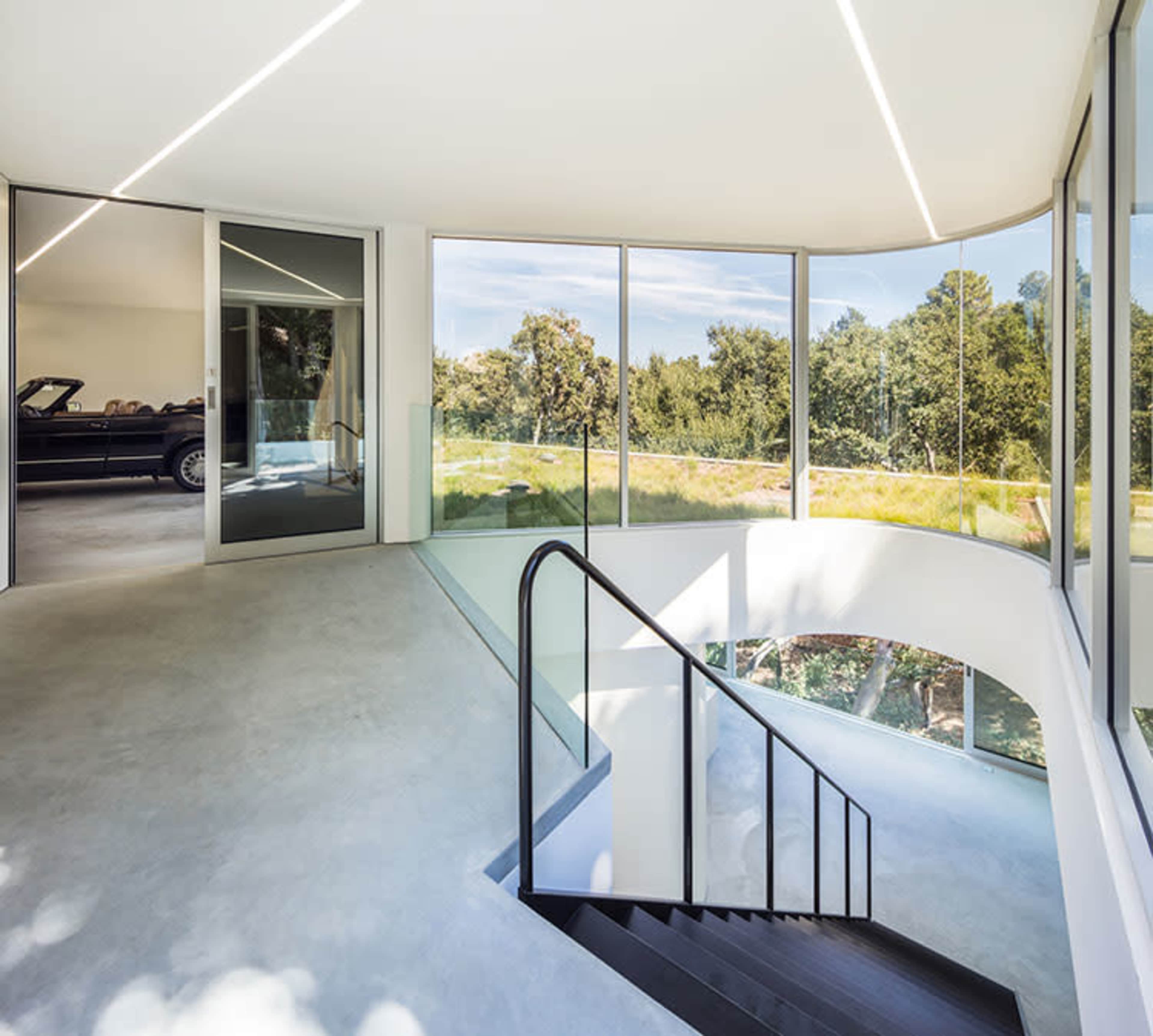 A modern interior space featuring a curved staircase, large glass windows, and a view of a car parked outside.