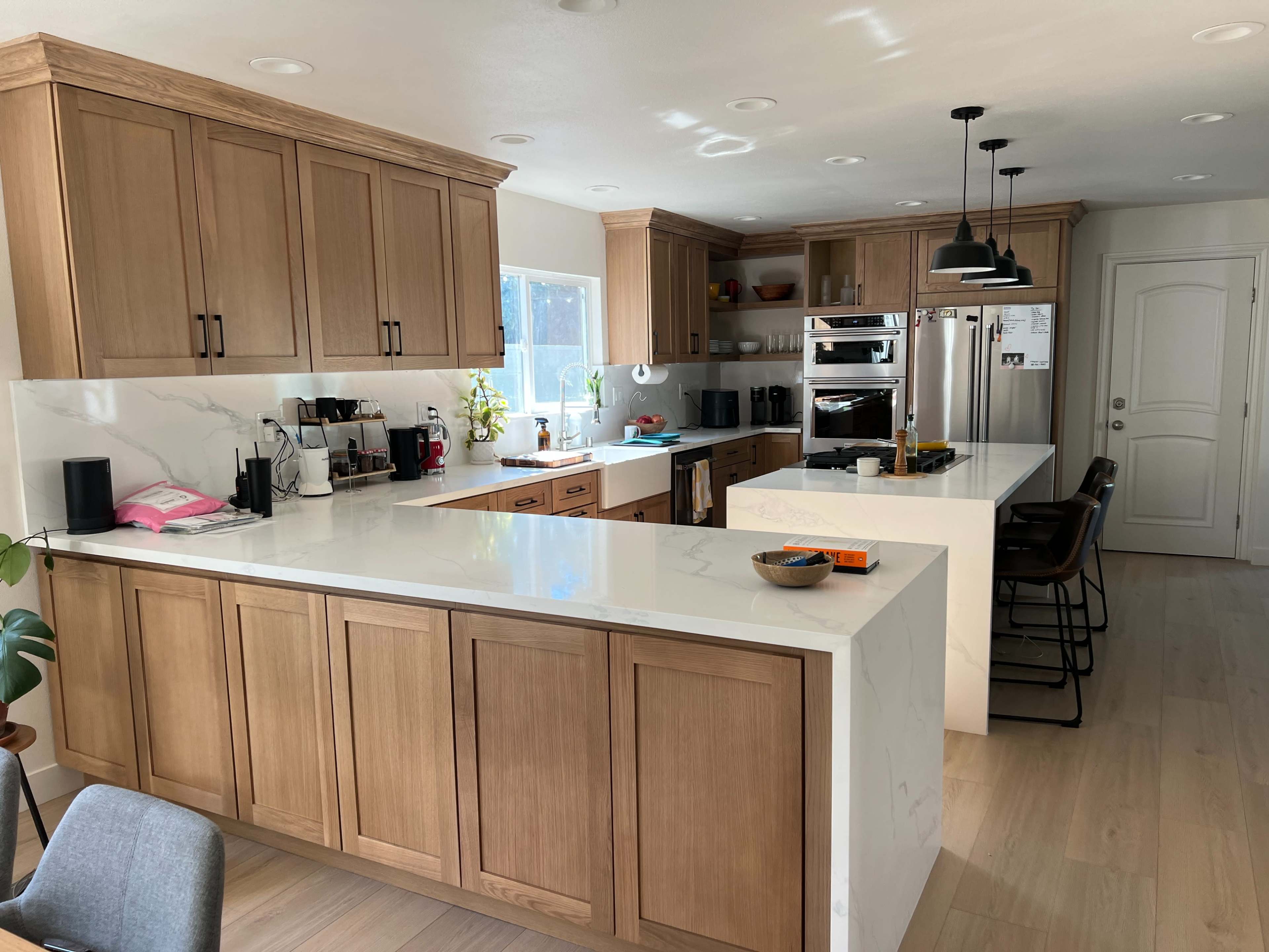 The image shows a modern kitchen with wood cabinets, a white marble countertop, and stainless steel appliances.