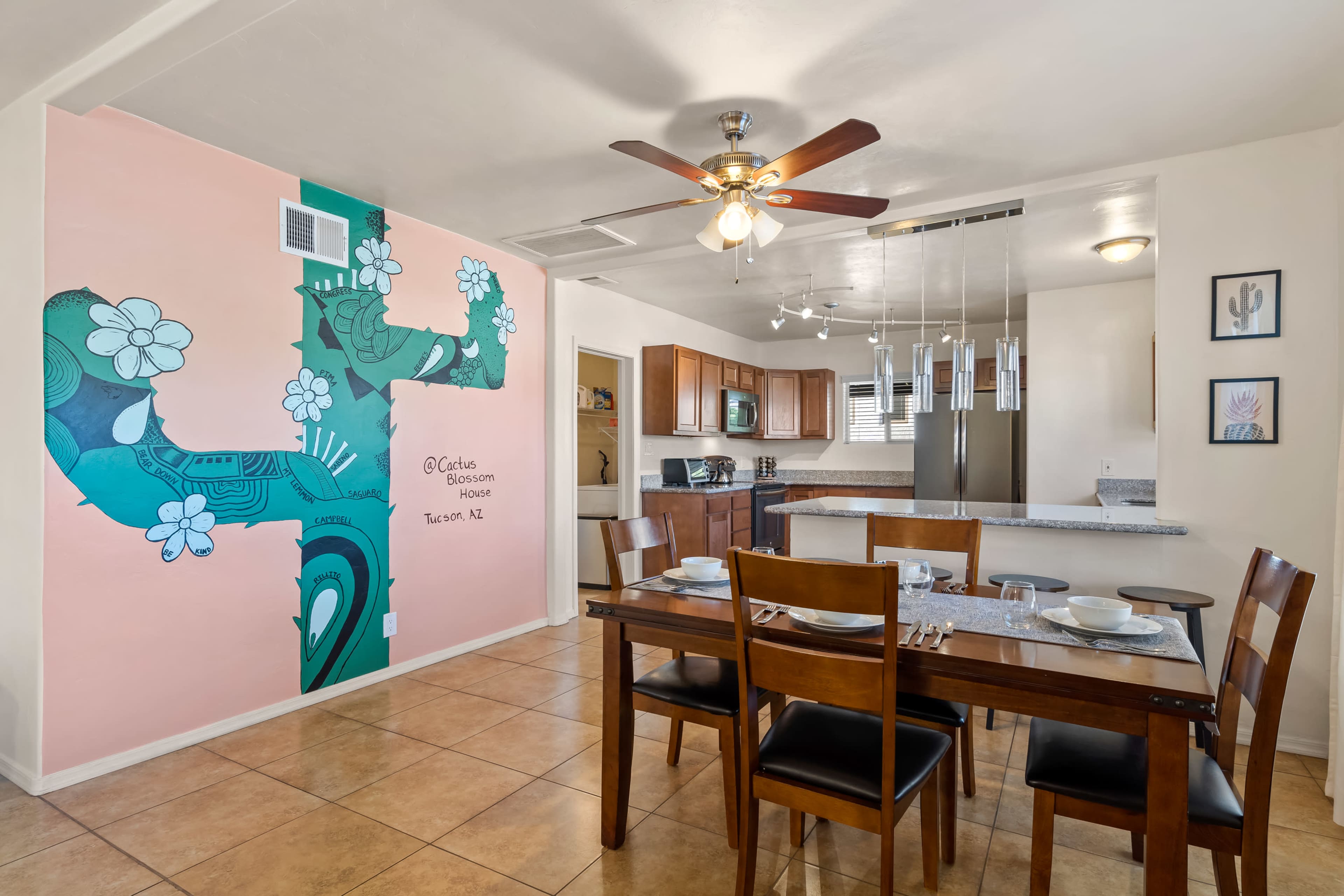 A dining area with a wooden table set for six is adjacent to a kitchen featuring warm wood cabinetry and a painted mural on the wall.