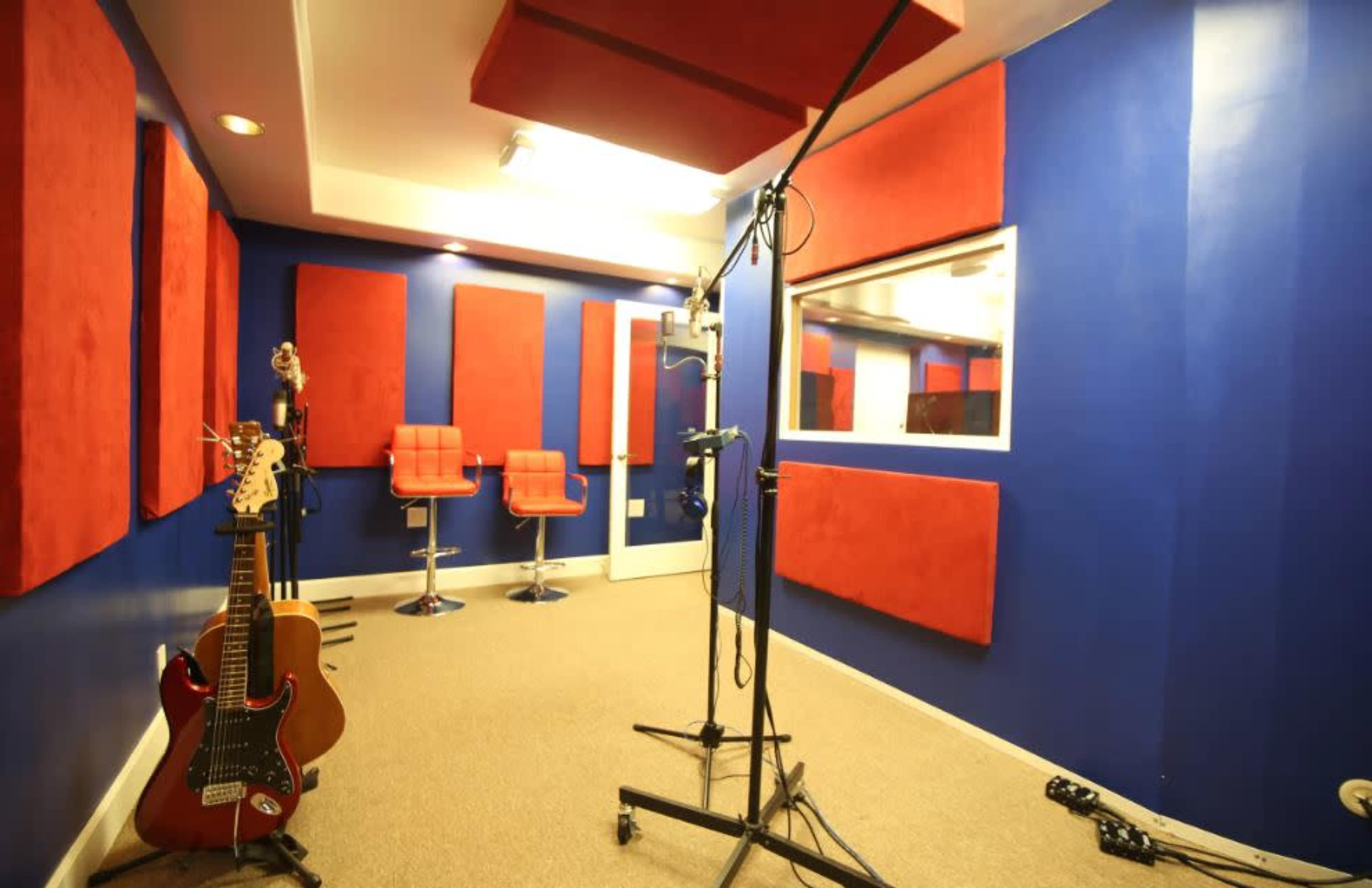 The image shows a sound studio with blue walls, red acoustic panels, a glass window, a microphone stand, and seating arrangements in the form of red stools.