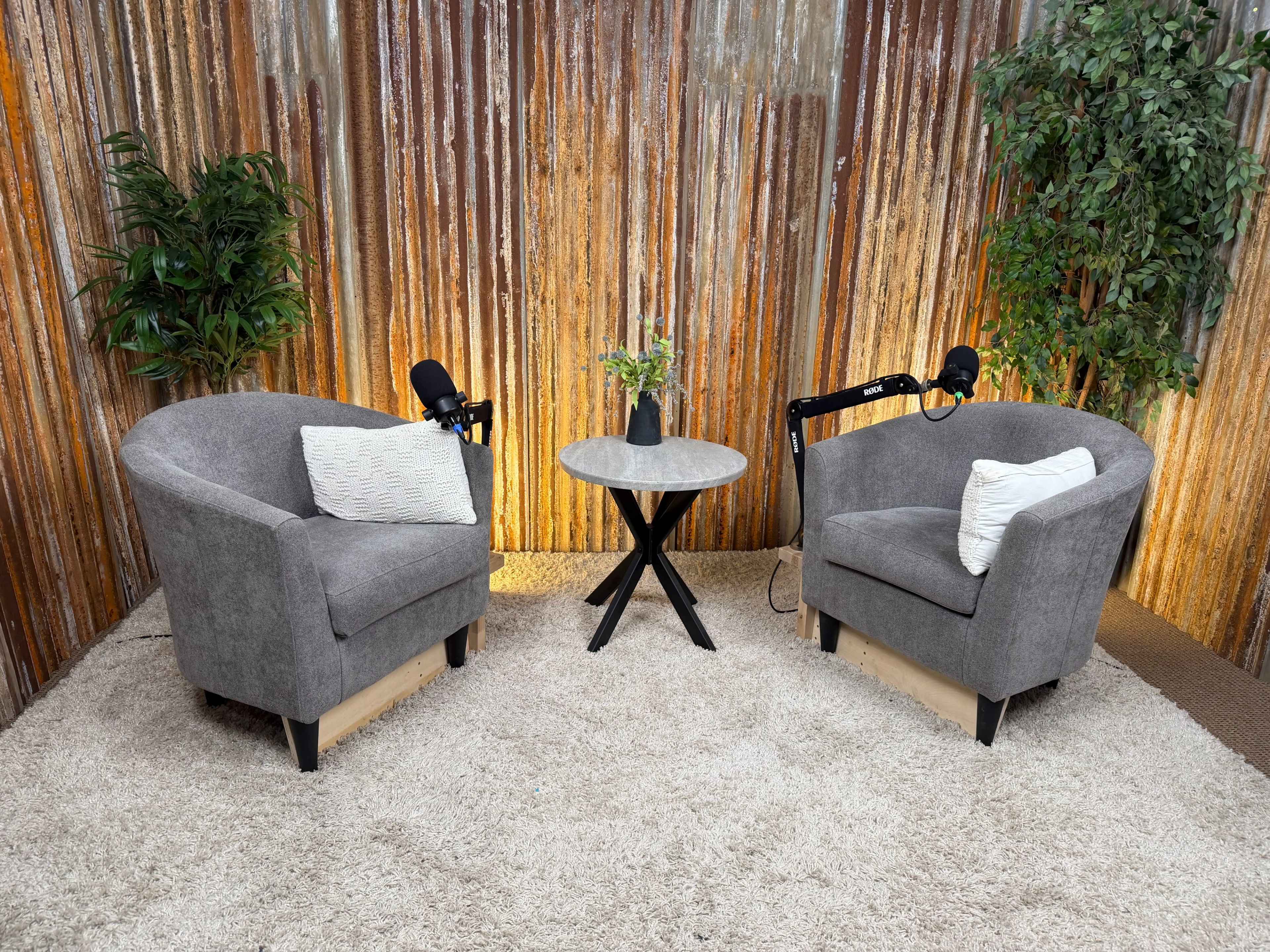 The image shows two cozy gray armchairs positioned opposite each other around a circular table, set against a rustic corrugated metal backdrop with soft lighting.