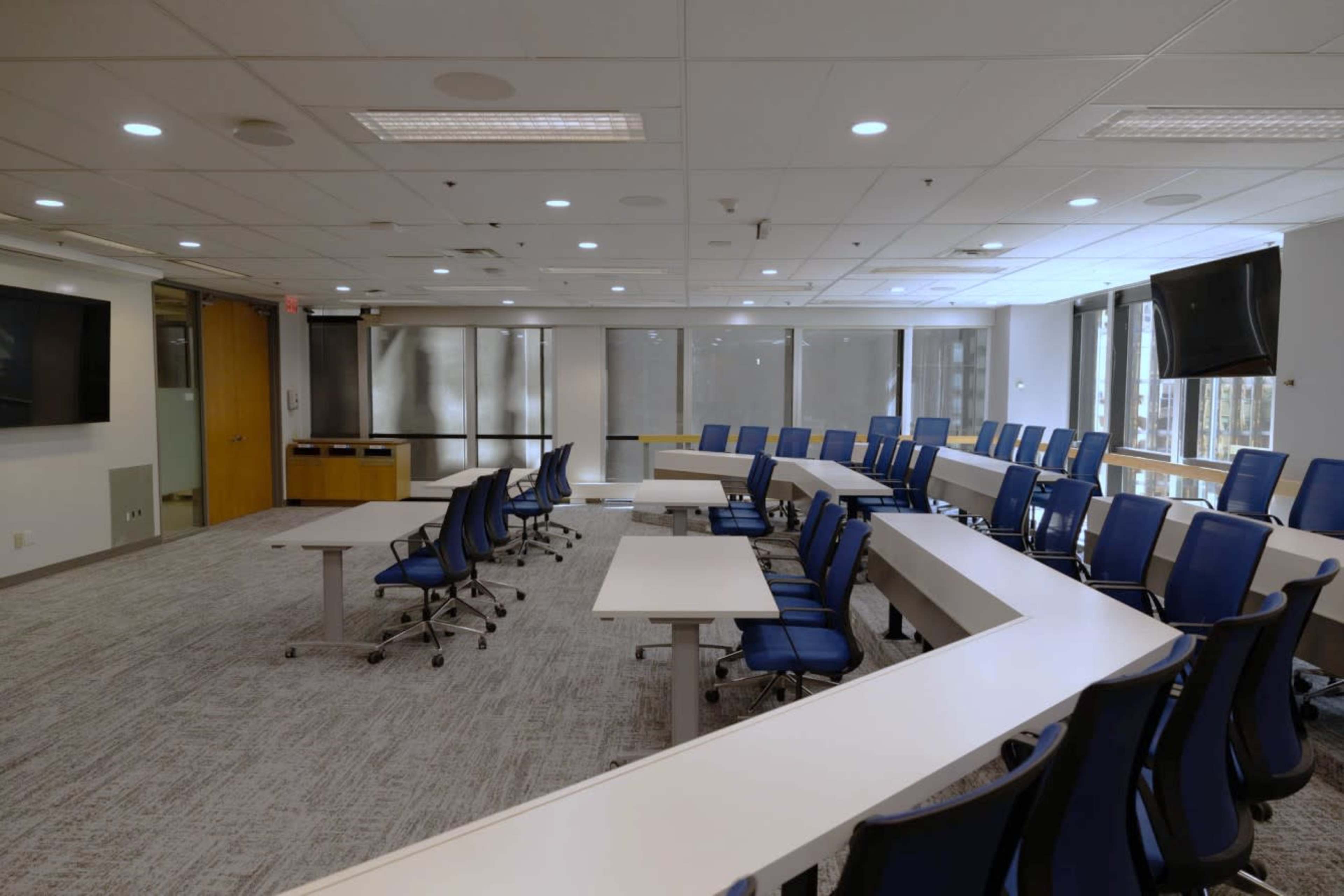 A modern conference room with a circular arrangement of white tables and blue chairs, equipped with large windows and two screens on the walls.
