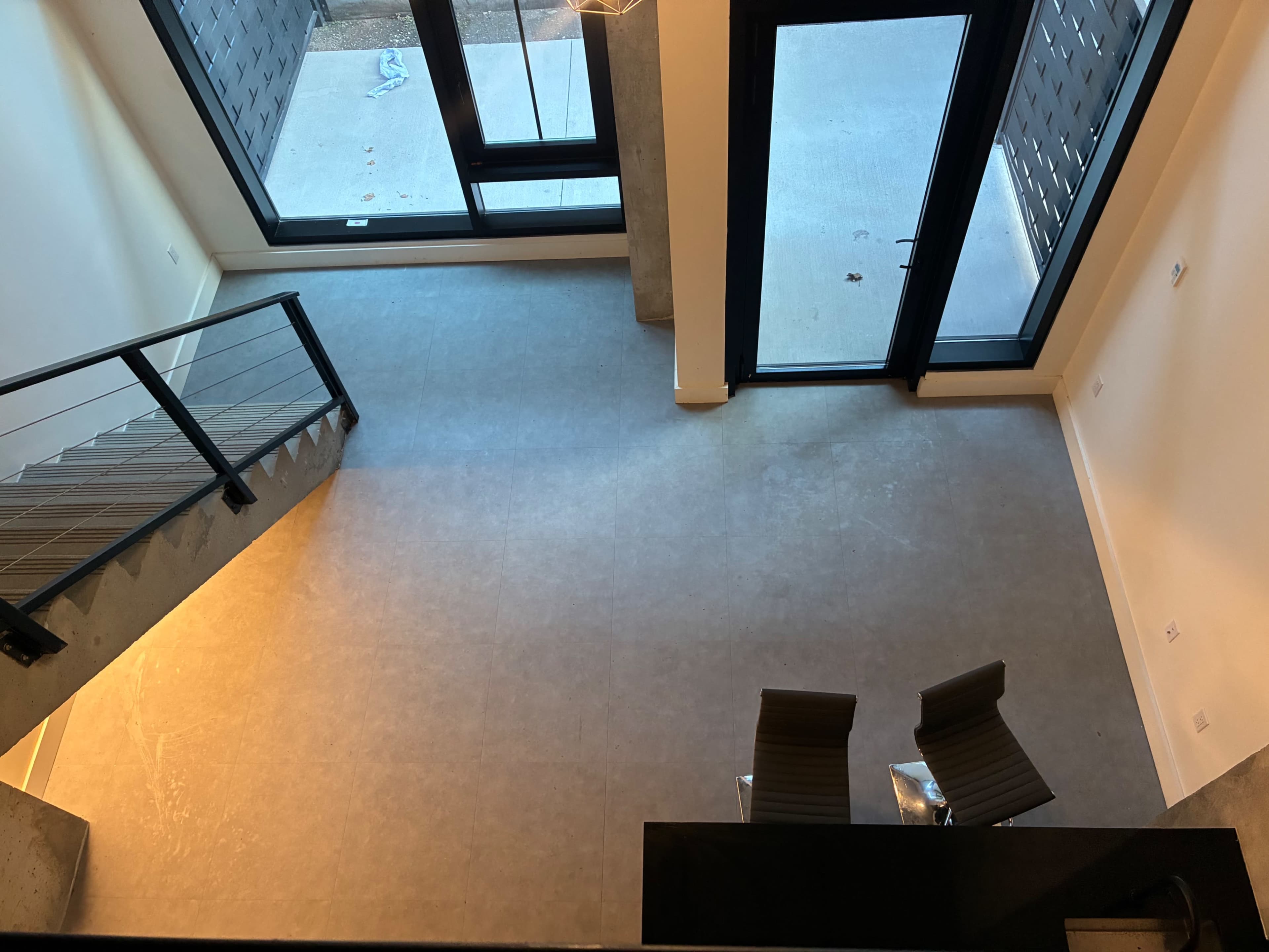The image shows a modern, open space with large windows, a concrete floor, and two chairs situated adjacent to a staircase.