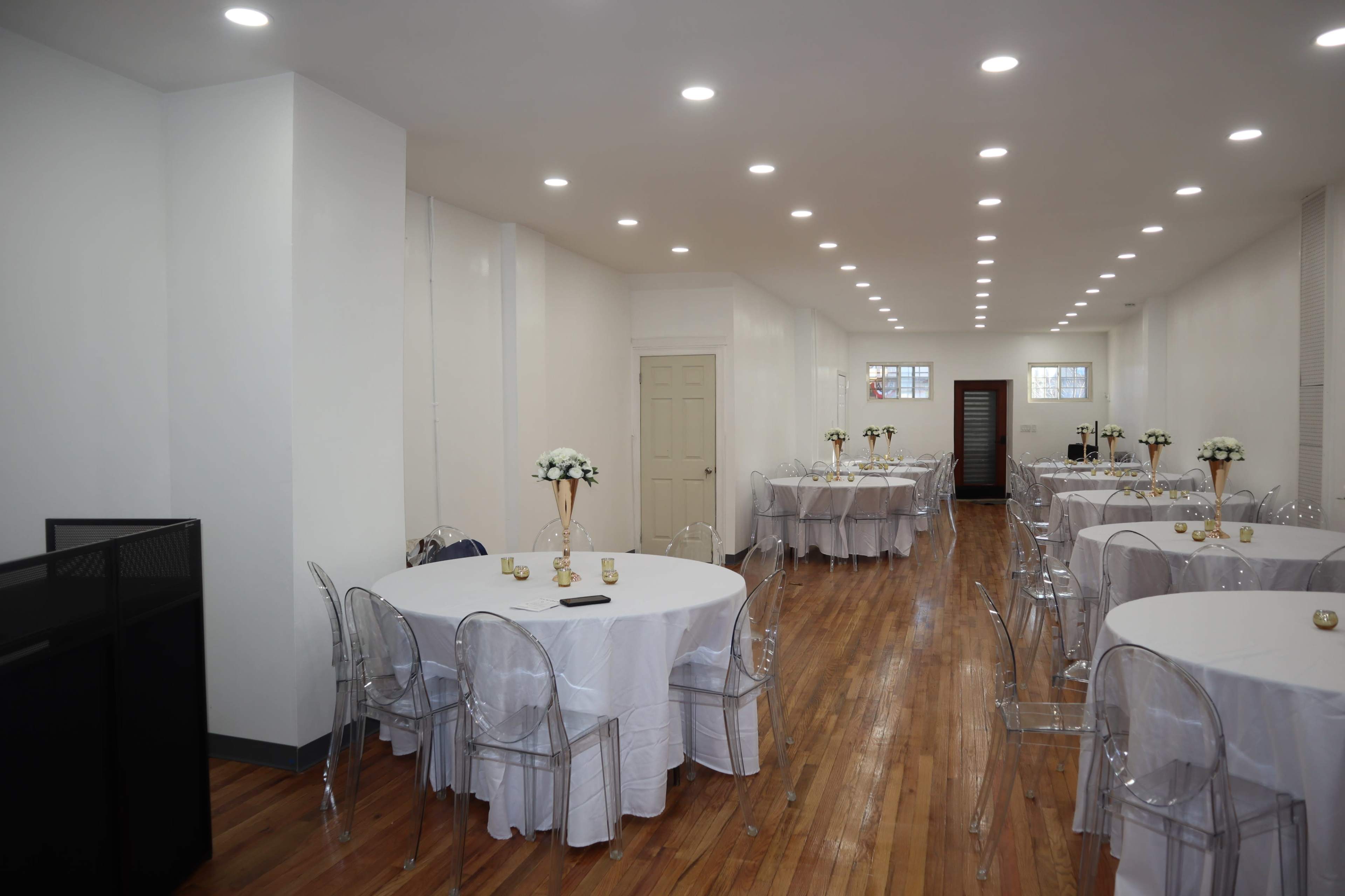 A spacious event room with multiple round tables covered in white tablecloths, clear chairs, and decorative centerpieces.