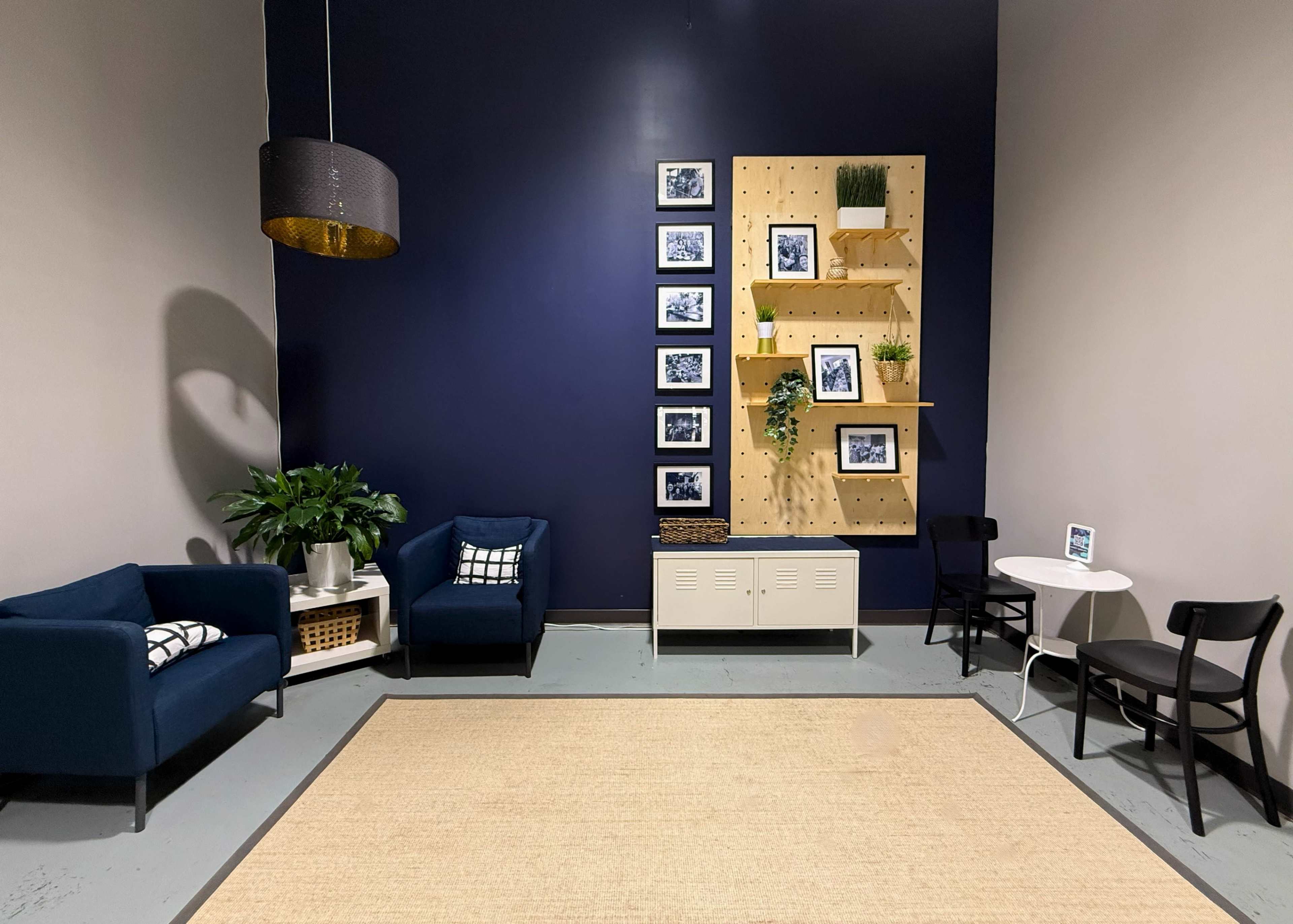 The image shows a small interior room featuring blue seating, a wooden wall shelf with framed pictures, and a light-colored rug on the floor.