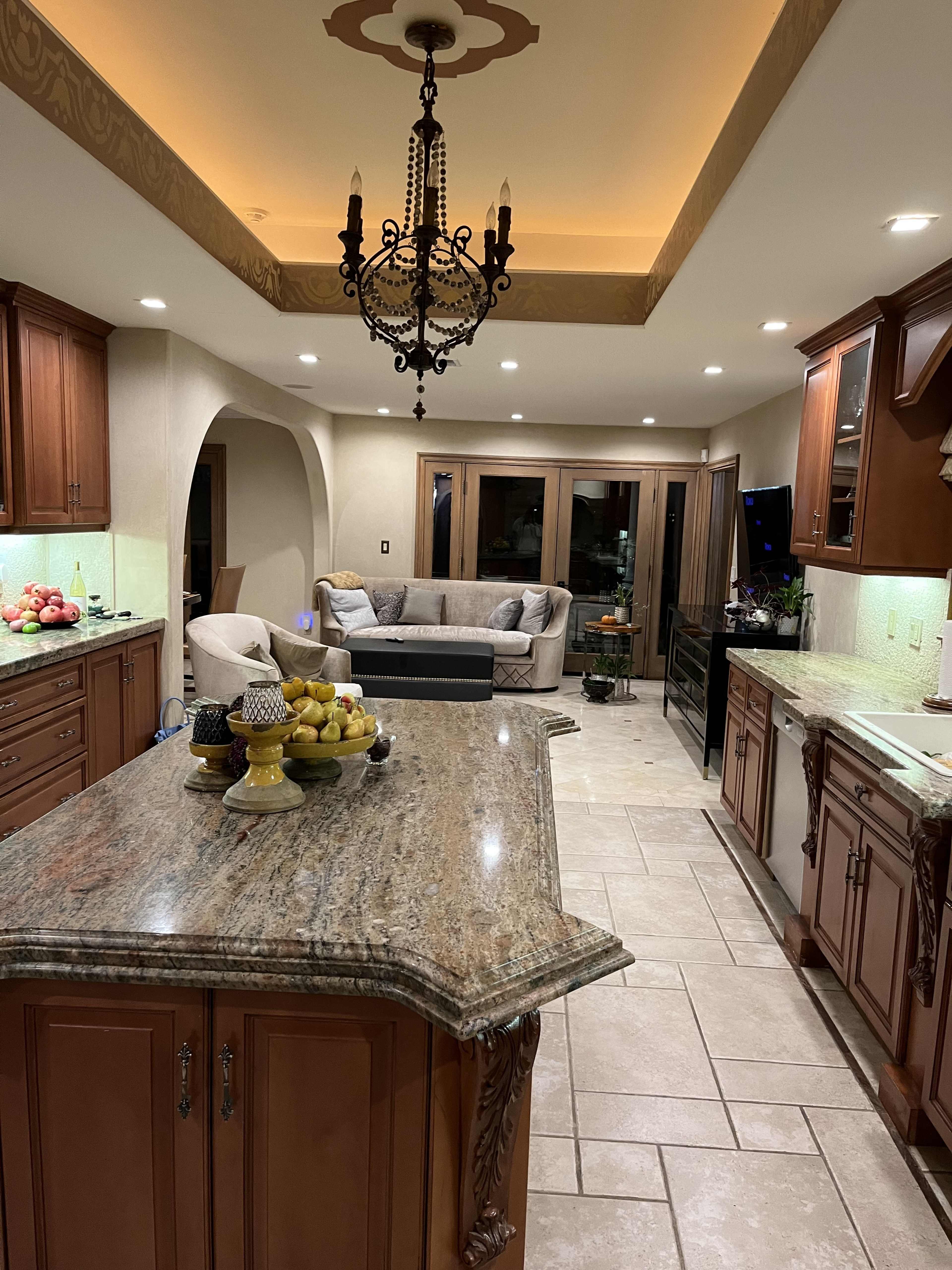 A spacious kitchen with dark wood cabinetry, a large granite island, and a living area featuring a light-colored sofa, illuminated by ceiling lights and a chandelier.