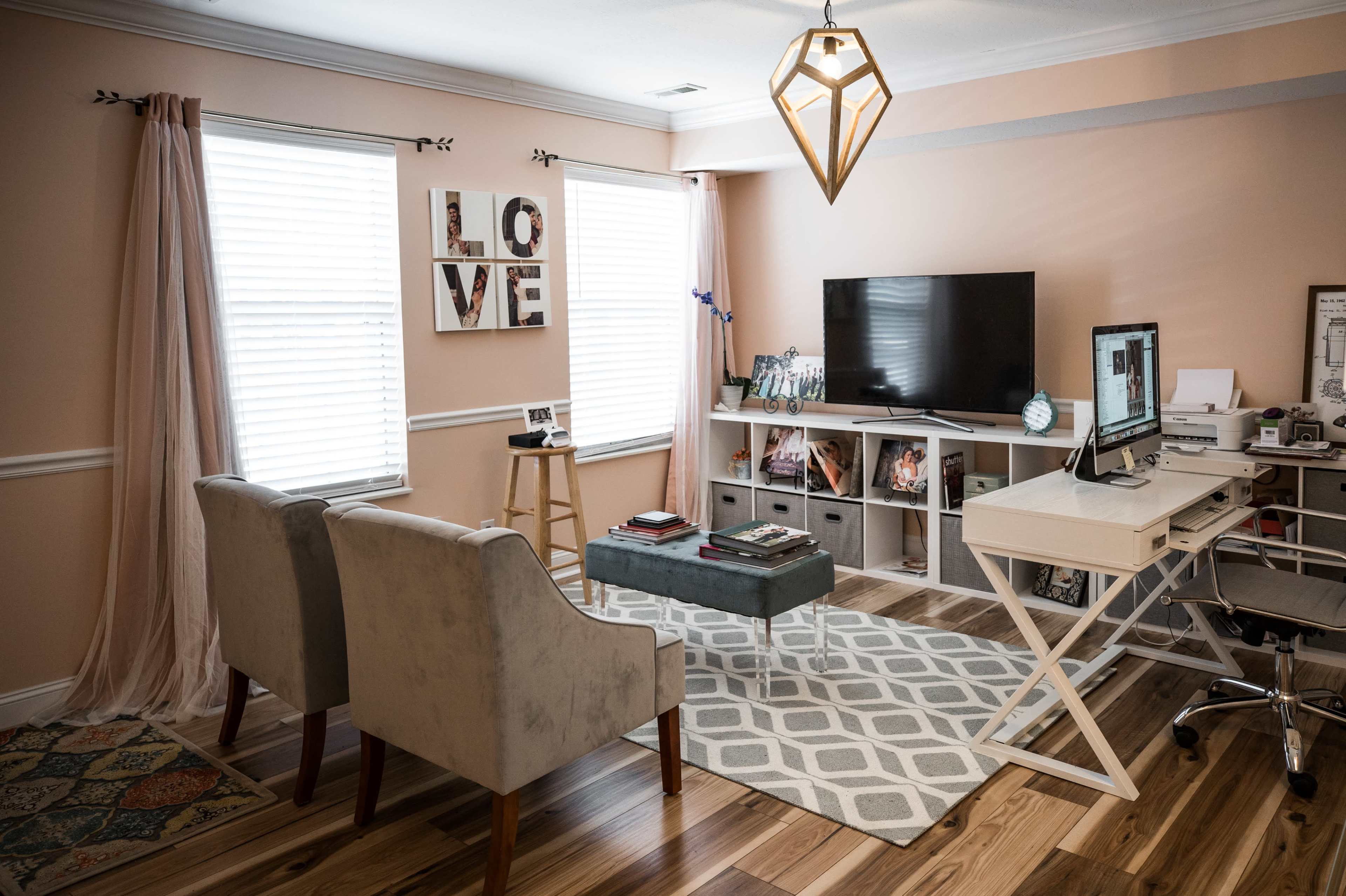 A modern living room features two gray chairs, a small green ottoman, a white desk with a computer, and a television mounted on a light-colored wall, with decorative elements including a "LOVE" sign and curtains by the windows.