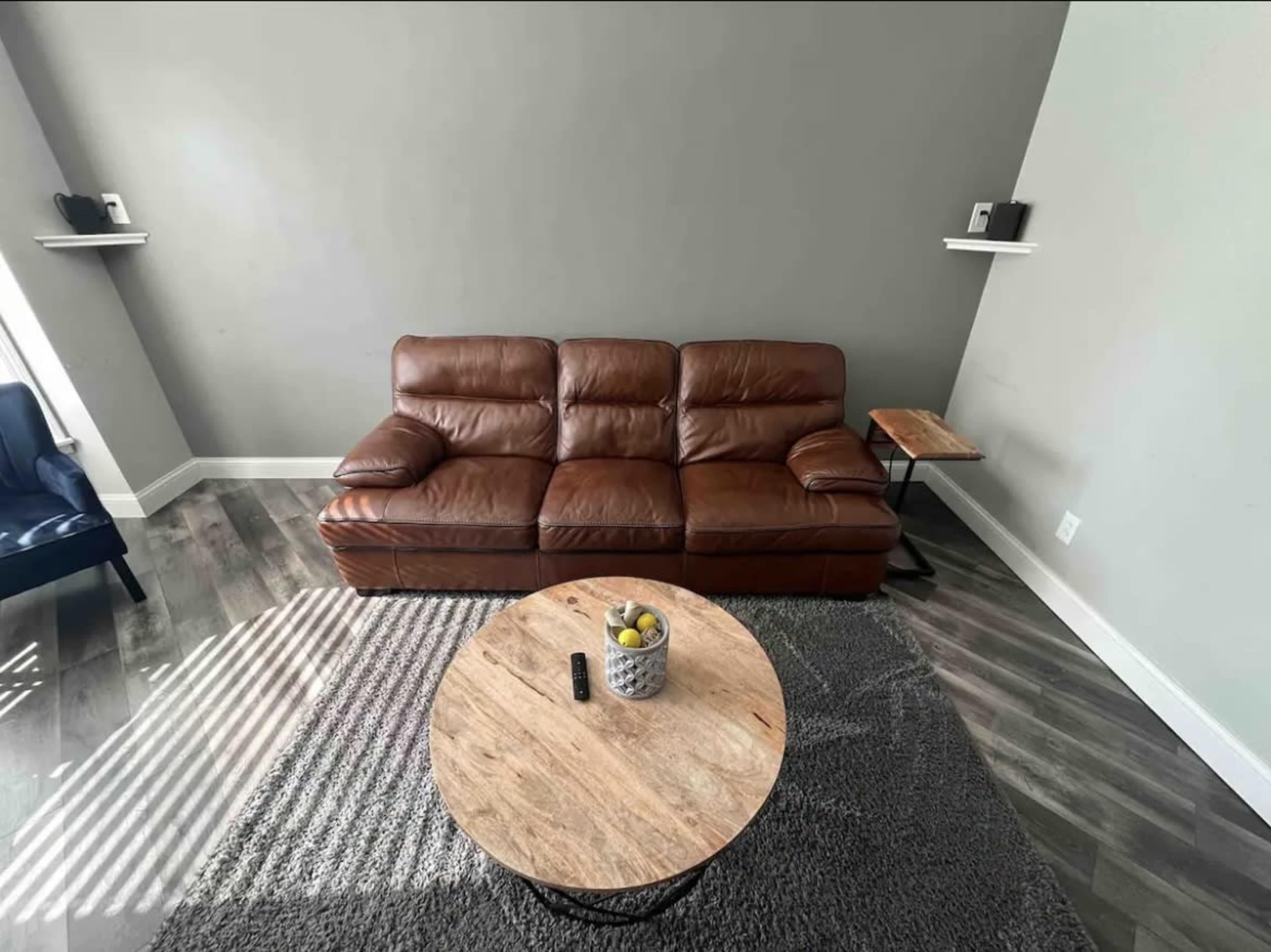A brown leather sofa sits in a minimalist living room with a round wooden coffee table and a textured gray rug.