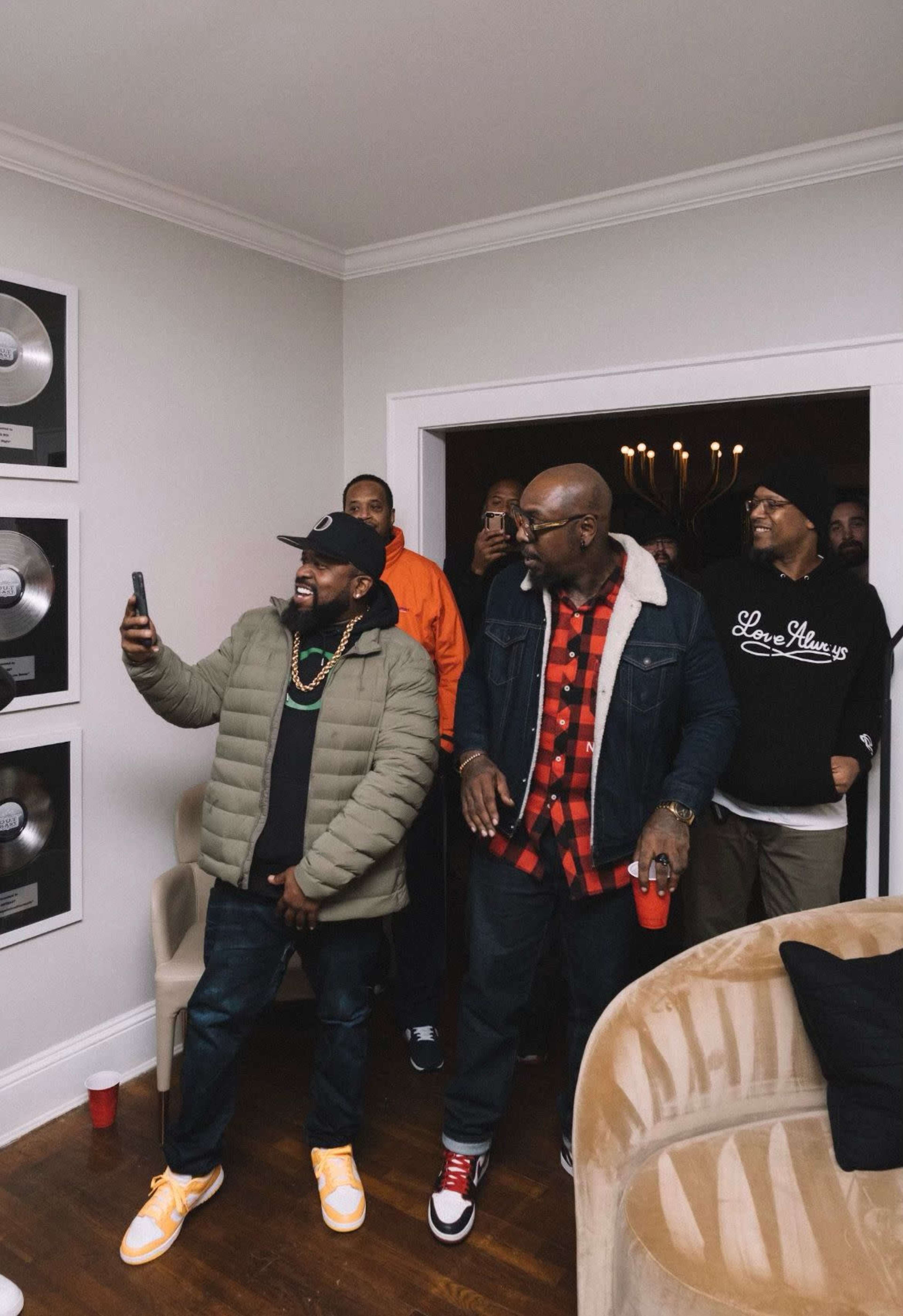 A group of men poses together in a living room, with one man checking his phone while others watch and smile.