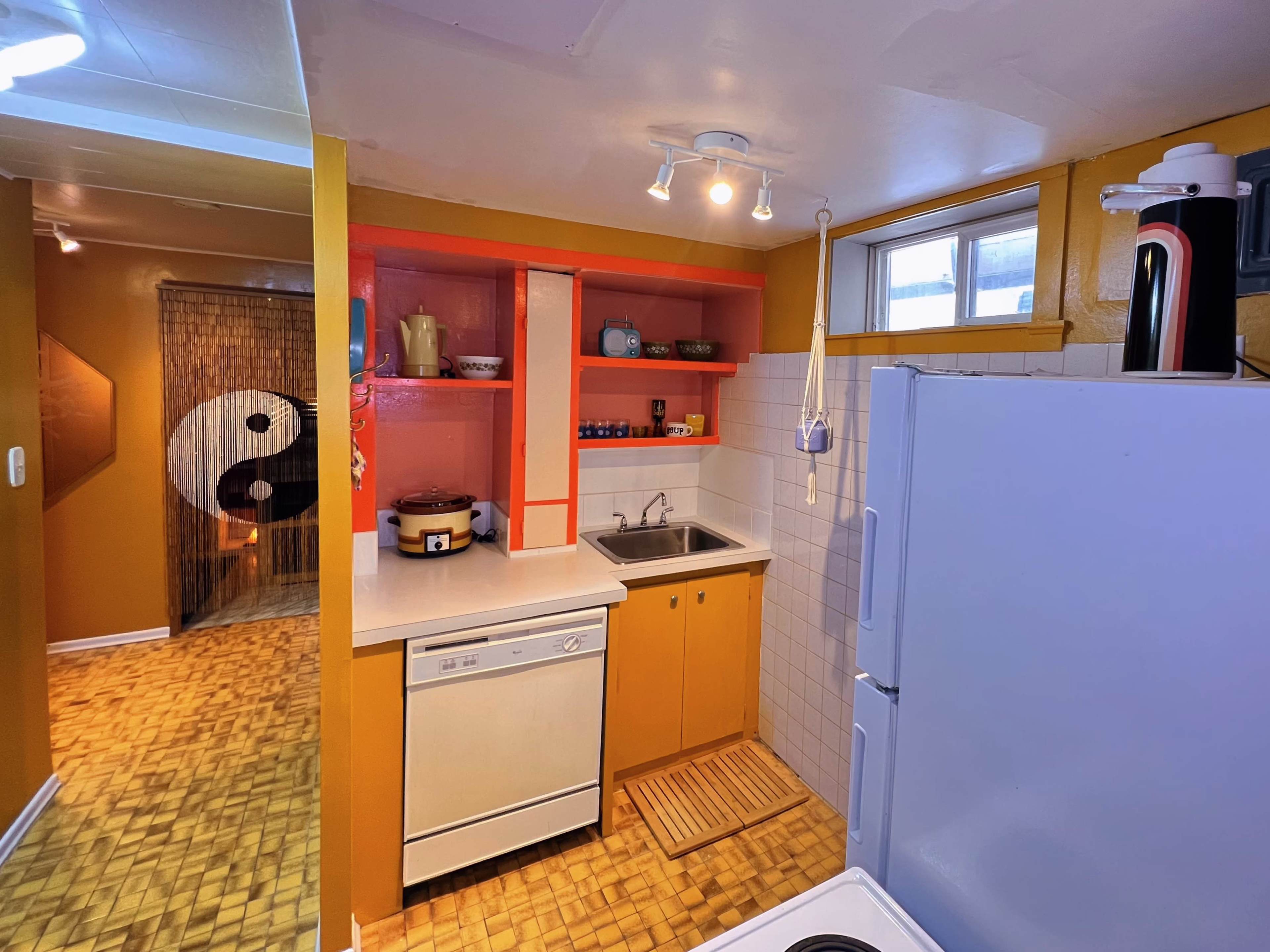 A small kitchen with orange and yellow walls, featuring a sink, dishwasher, and open shelving, leading into a room with a decorative yin-yang design.