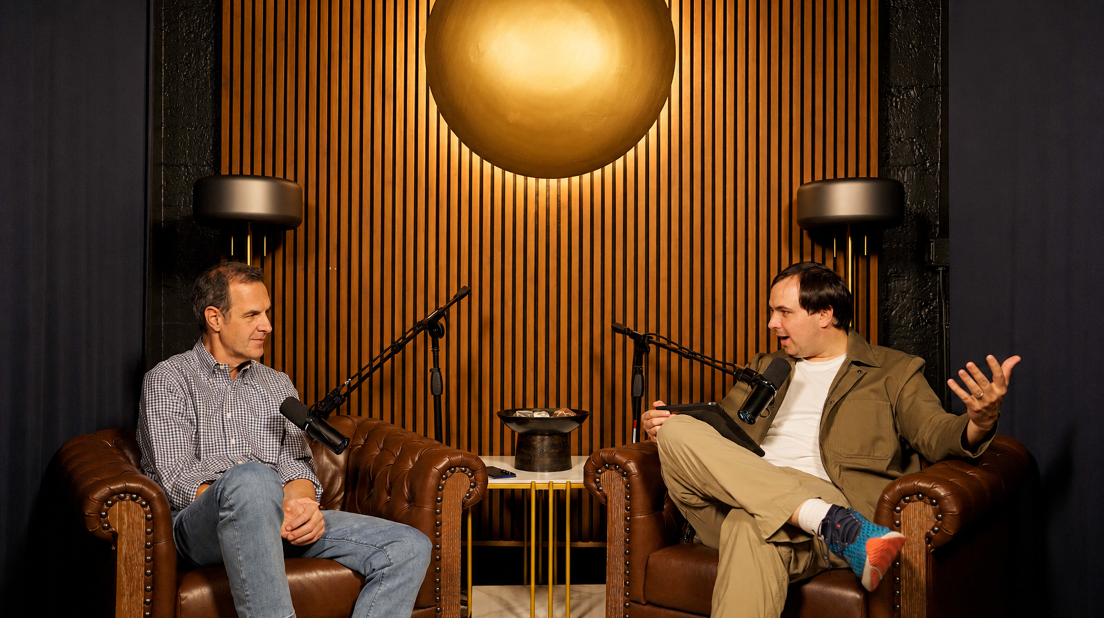 Two men engage in conversation while seated in a modern studio with a golden circular light fixture and wood-paneled wall behind them.
