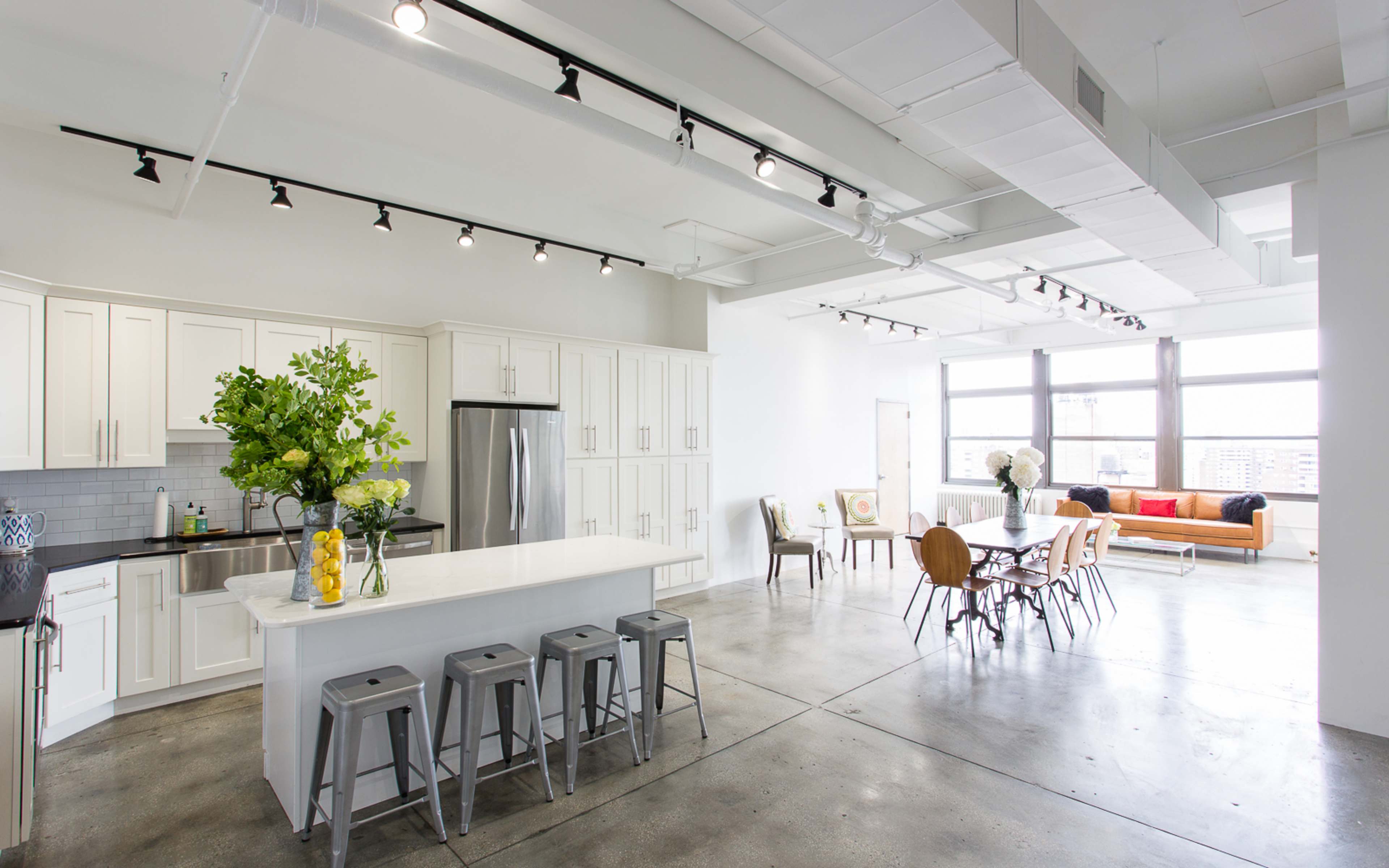 A modern open-concept kitchen and dining area with white cabinetry, a large island, and a dining table surrounded by chairs, all set in a well-lit space with industrial-style concrete flooring.
