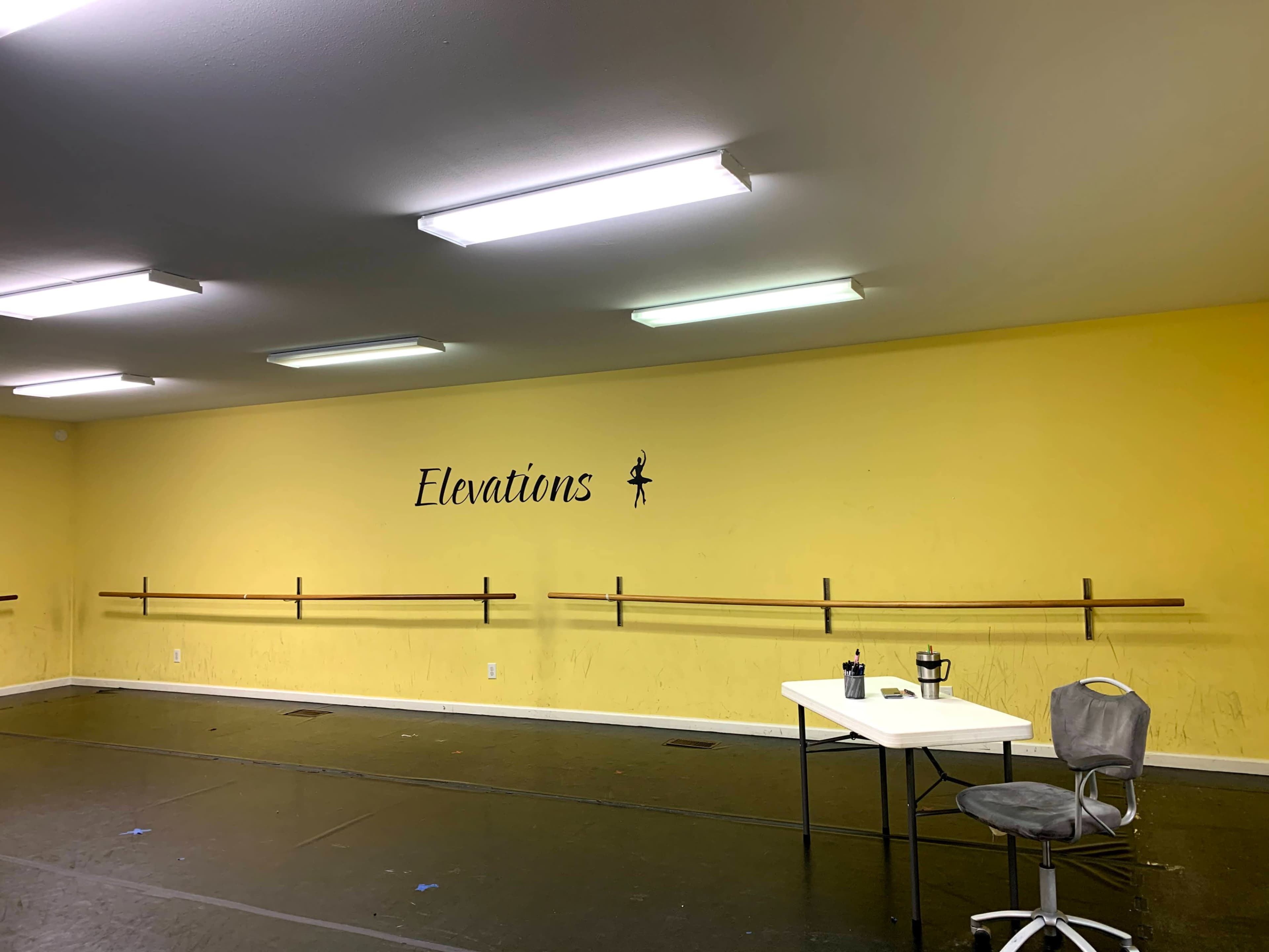 A bright yellow dance studio features a barre along the wall and a table with a chair.