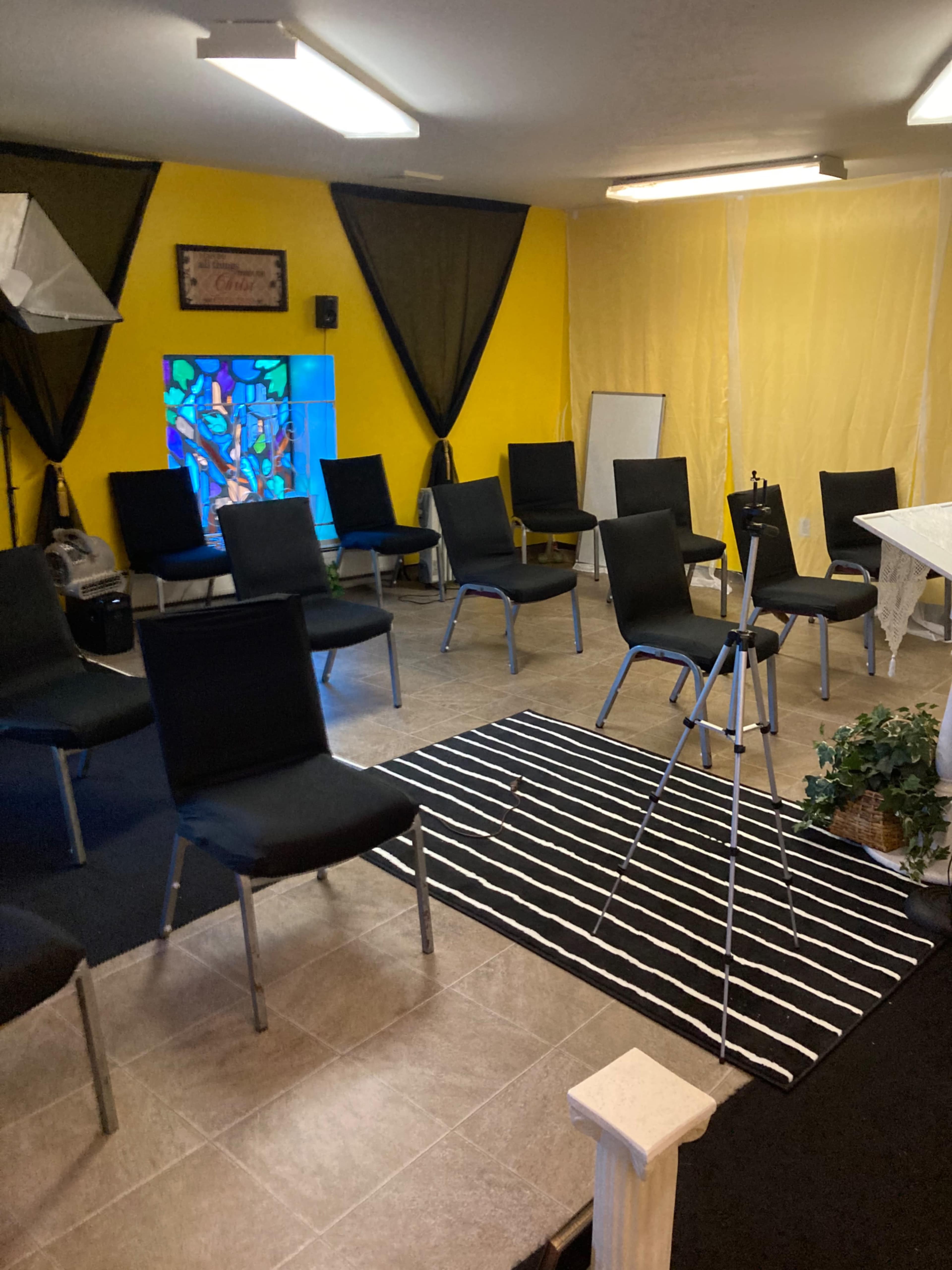 A room set up with black chairs arranged in a circular formation around a tripod and a small table, featuring yellow walls and a stained glass window.