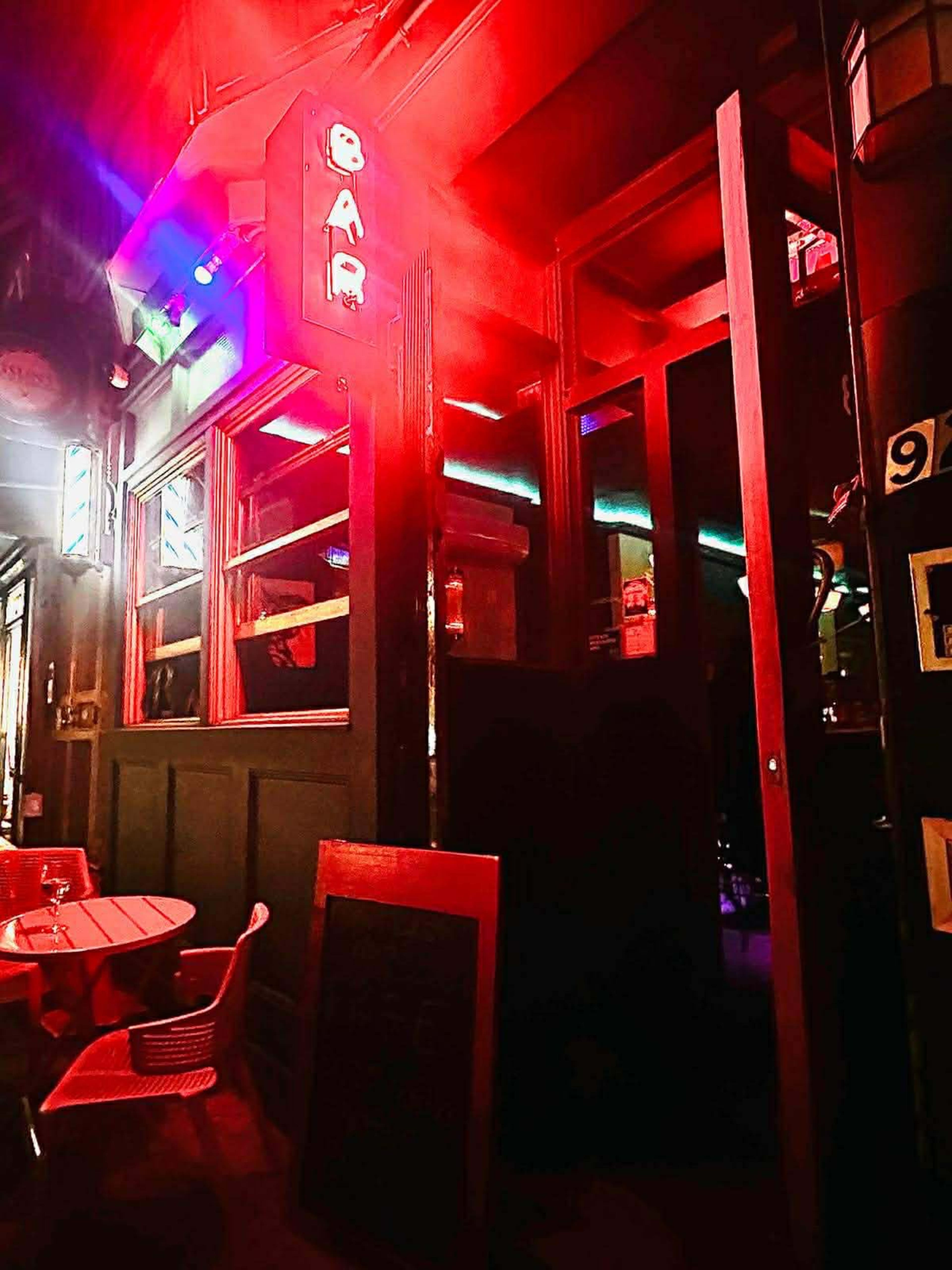 A dimly lit bar entrance with red neon lights and a small outdoor seating area.