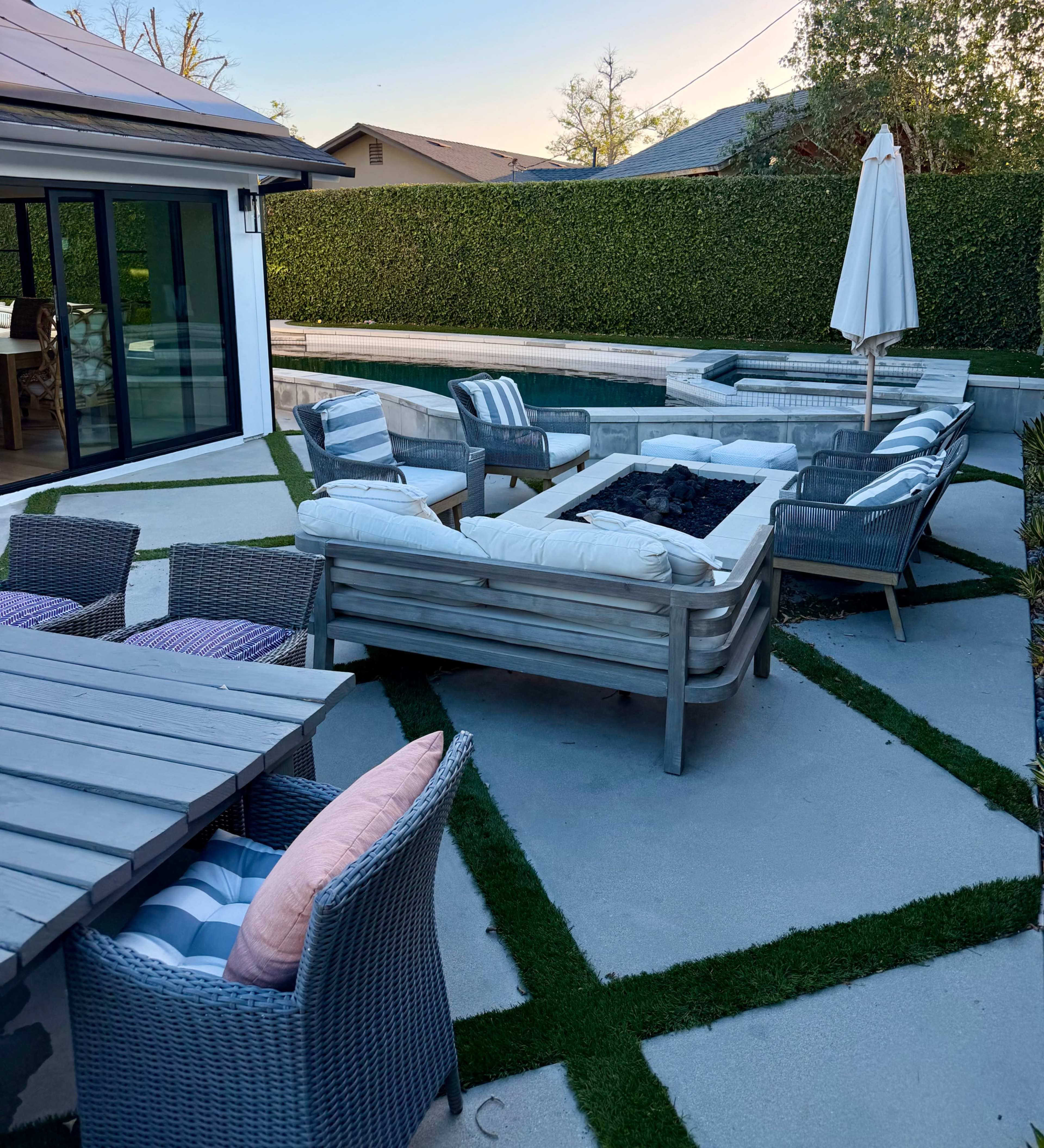 A patio area features modern outdoor furniture arranged around a fire pit, with artificial grass accents and a hedge backdrop.