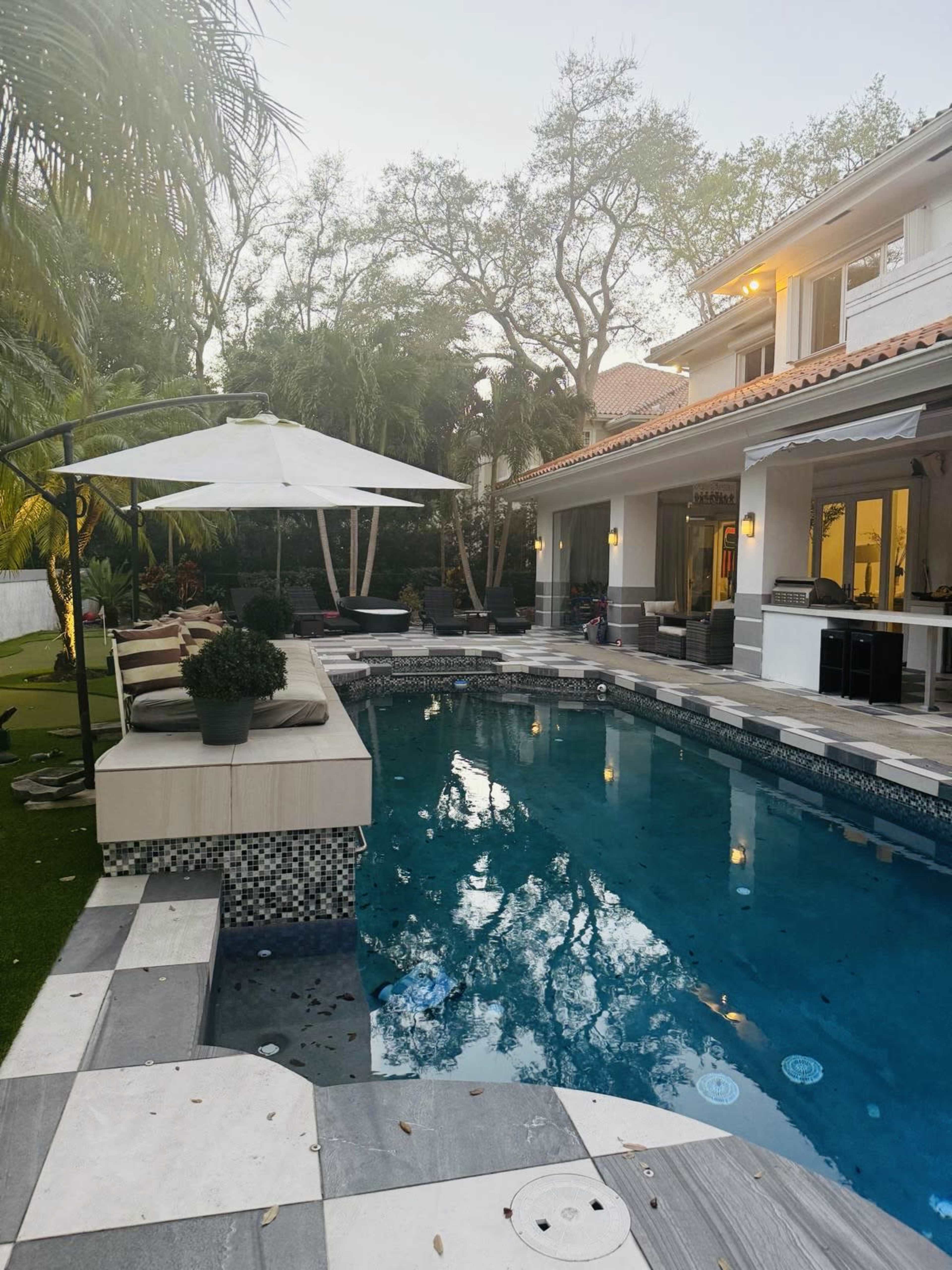 A modern backyard features a swimming pool surrounded by patio tiles, a shaded seating area, and lush greenery.