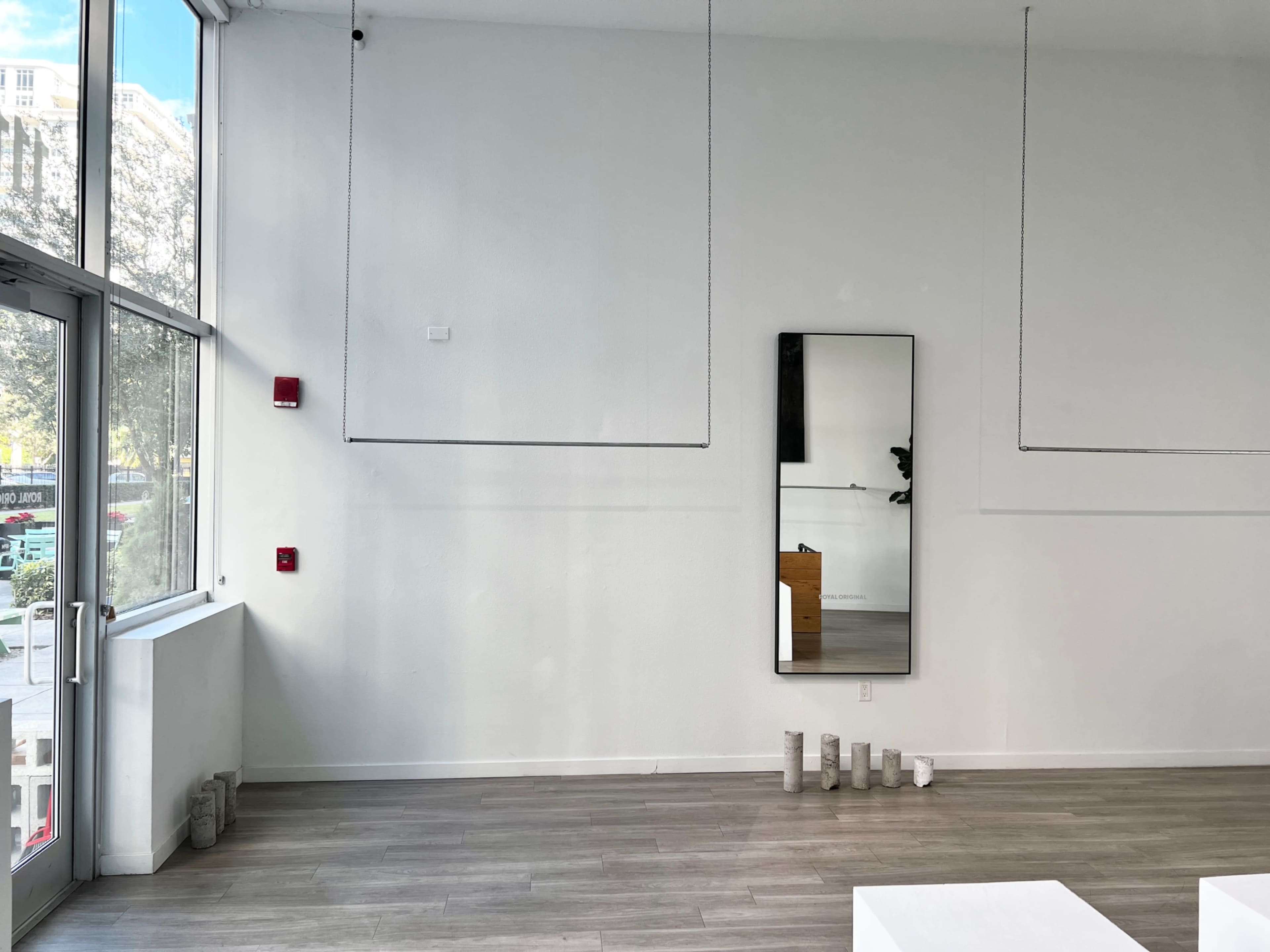 A minimalist interior space with a large mirror on the wall, a few light-colored rectangular objects on the floor, and a window letting in natural light.