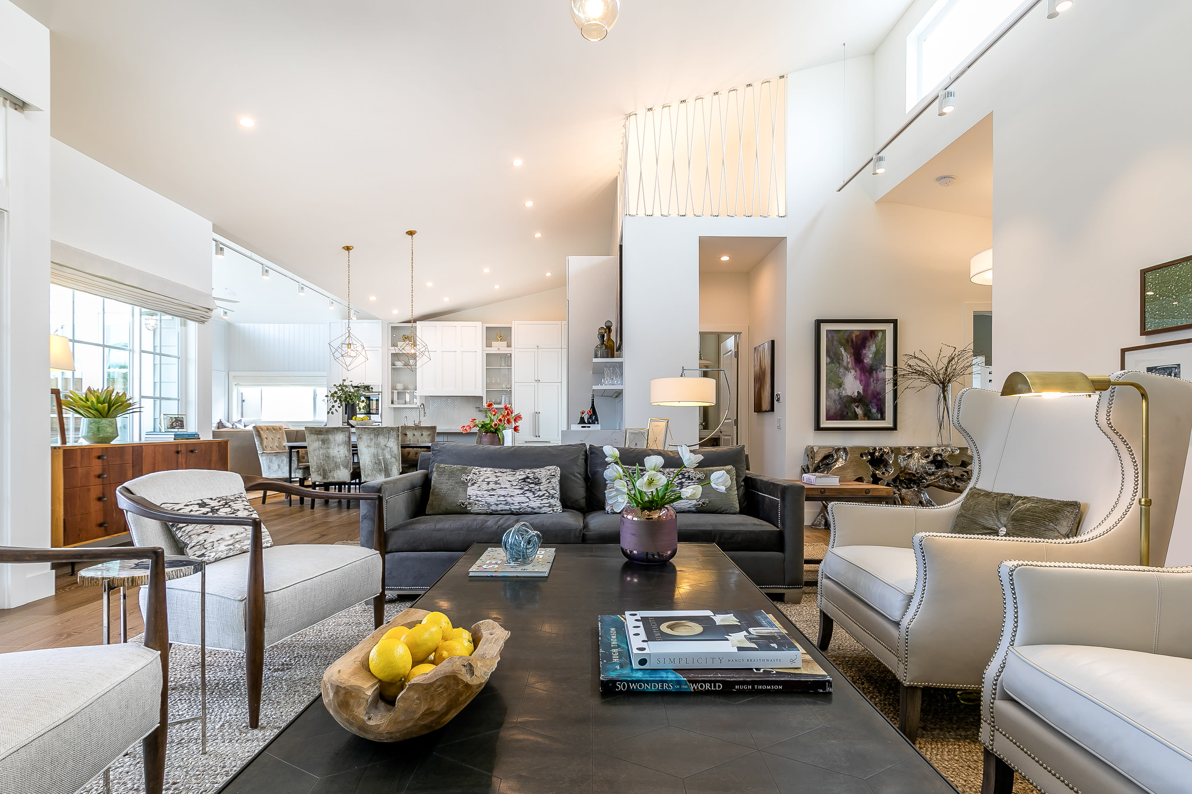 A modern living room with a dark sofa, two armchairs, and a coffee table adorned with decor and a bowl of lemons, connected to a sleek kitchen in the background.