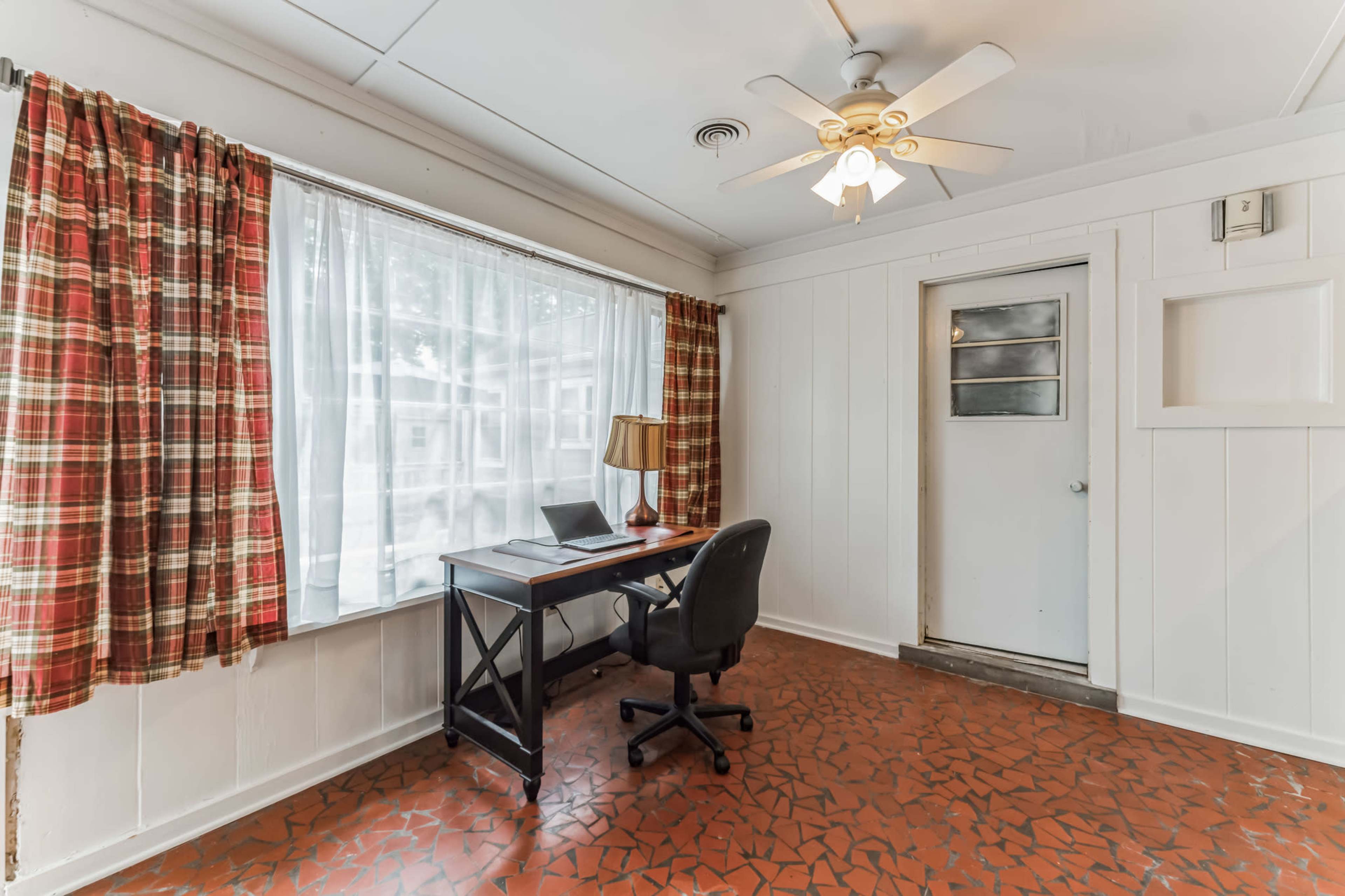 The image shows a small room with a desk and a chair, a window with checkered curtains, and a door leading outside.