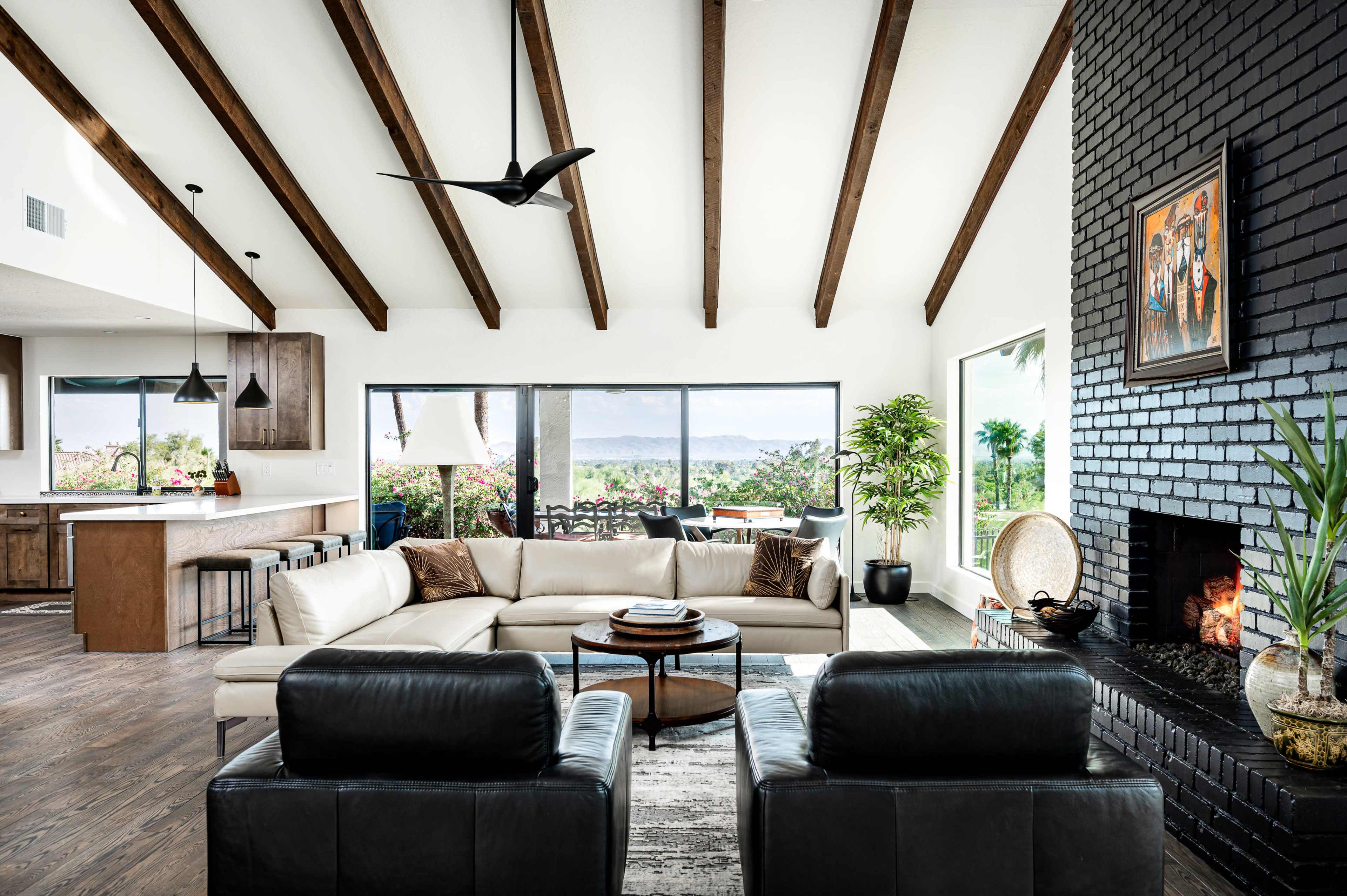 A modern living room with a high ceiling, featuring a sectional sofa, two black chairs, a stone fireplace, and large windows overlooking a garden.