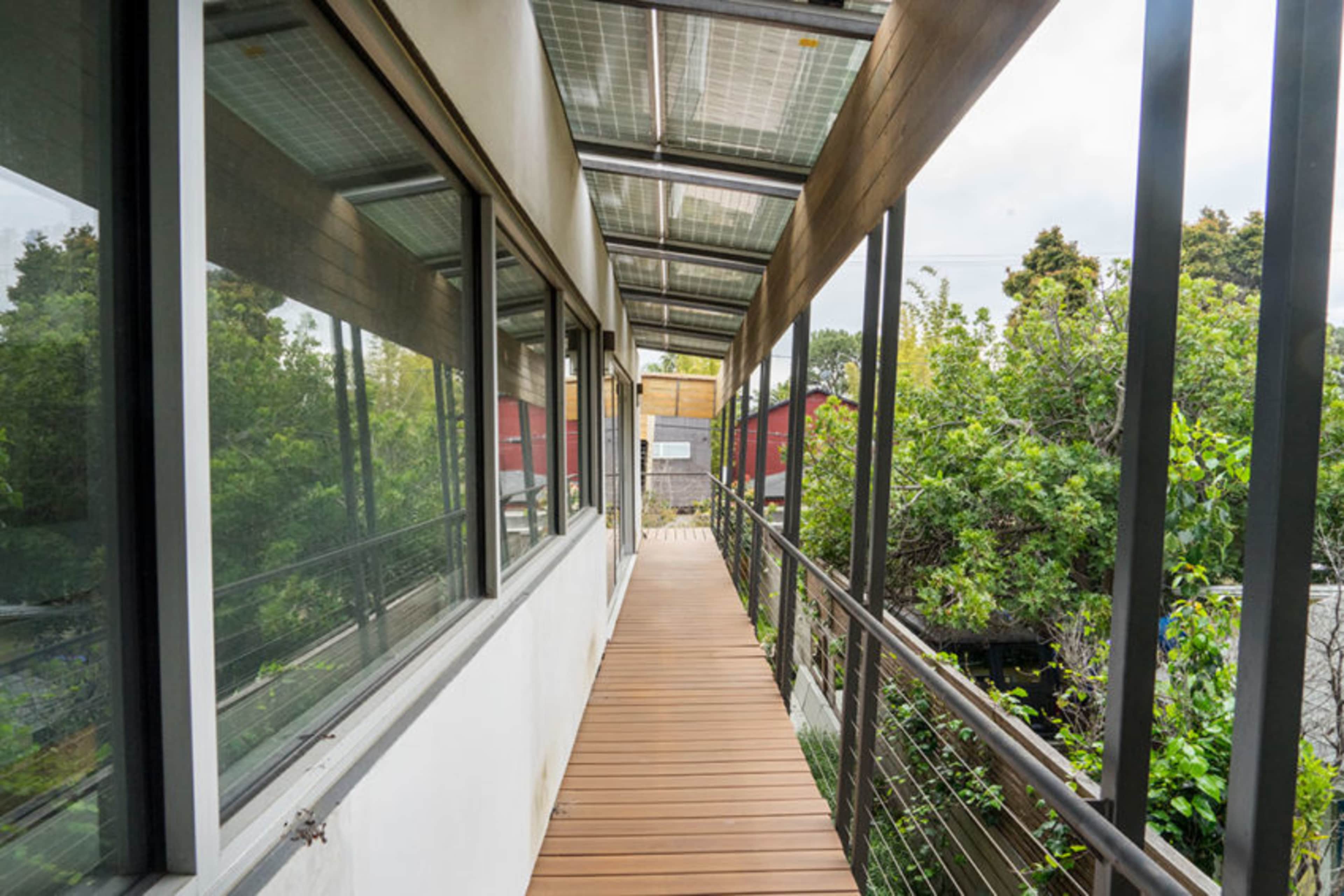 The image depicts a wooden walkway enclosed by large windows, leading to a green, tree-filled area.