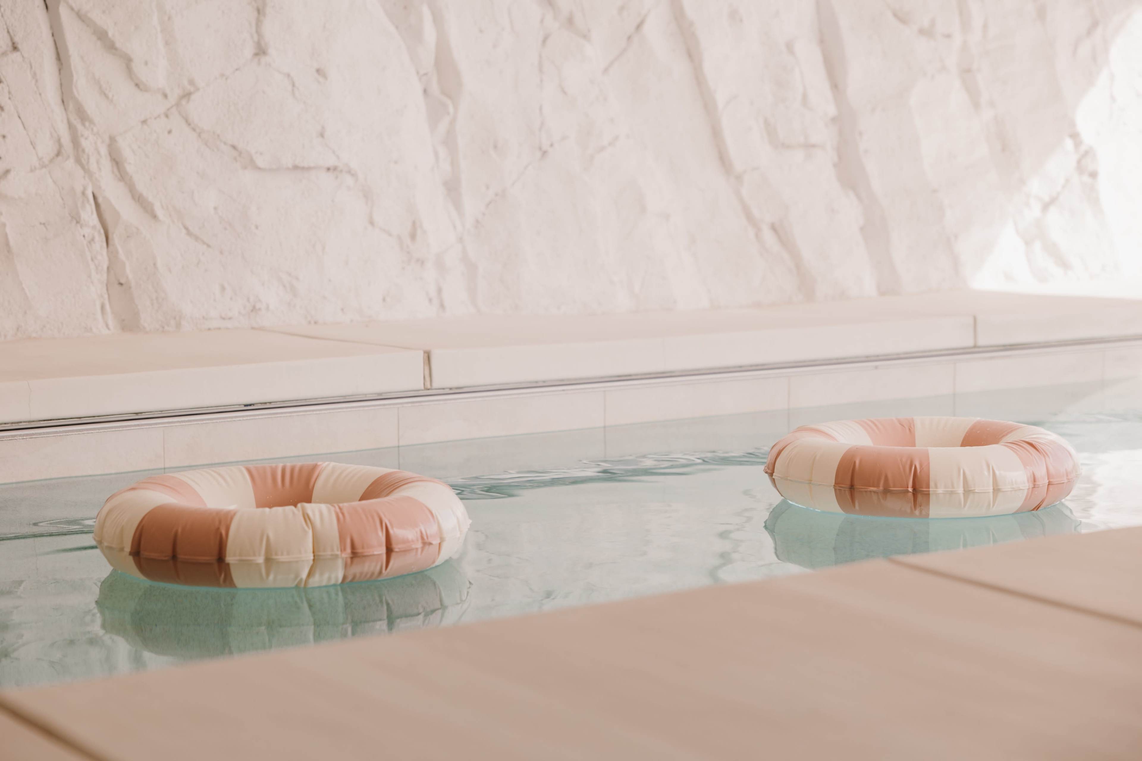 Two pastel-colored pool floats are set in still water beside a textured white stone wall.