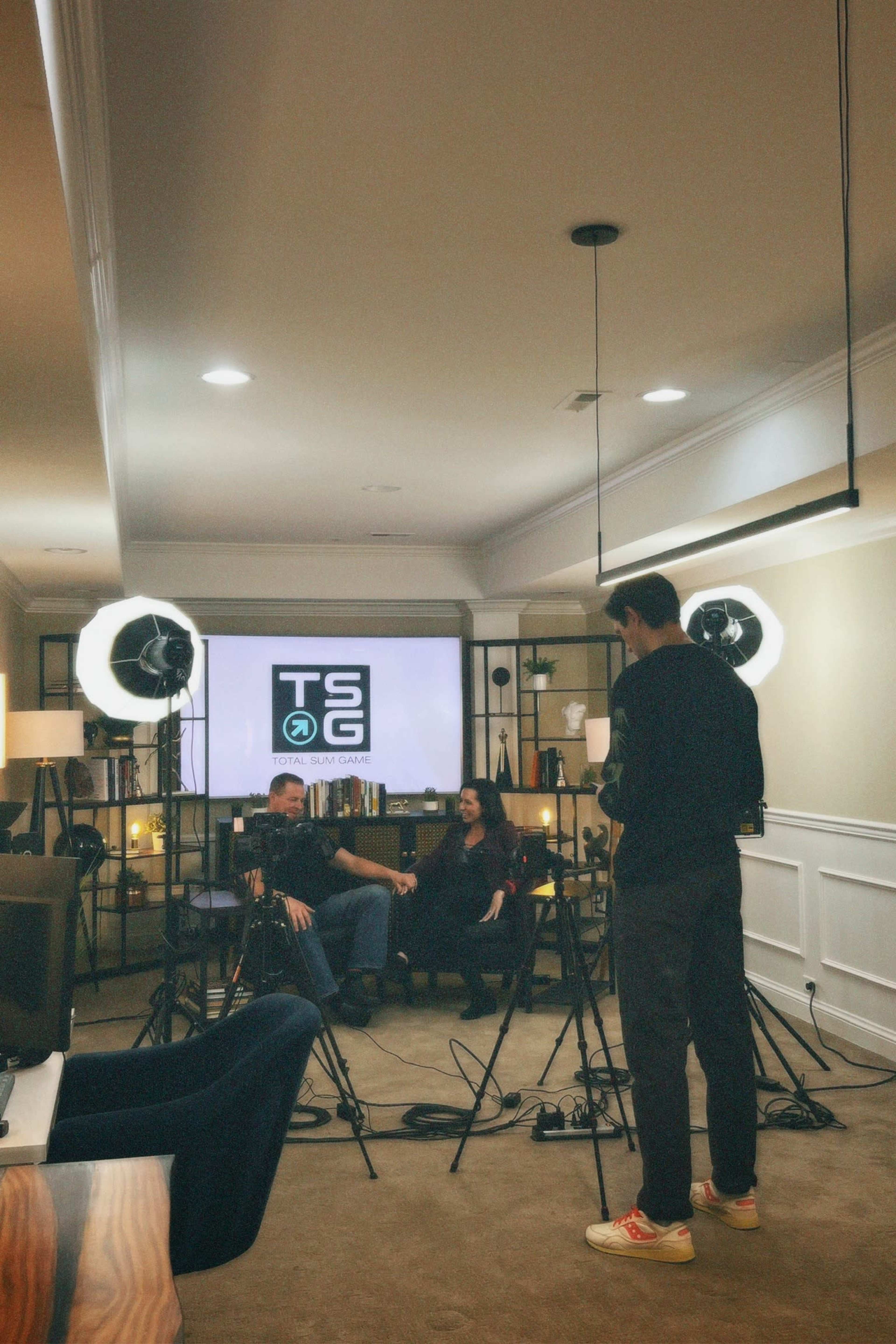 A man and a woman are seated in a well-lit studio space, with cameras and lights set up in front of a backdrop featuring the logo "TSG."