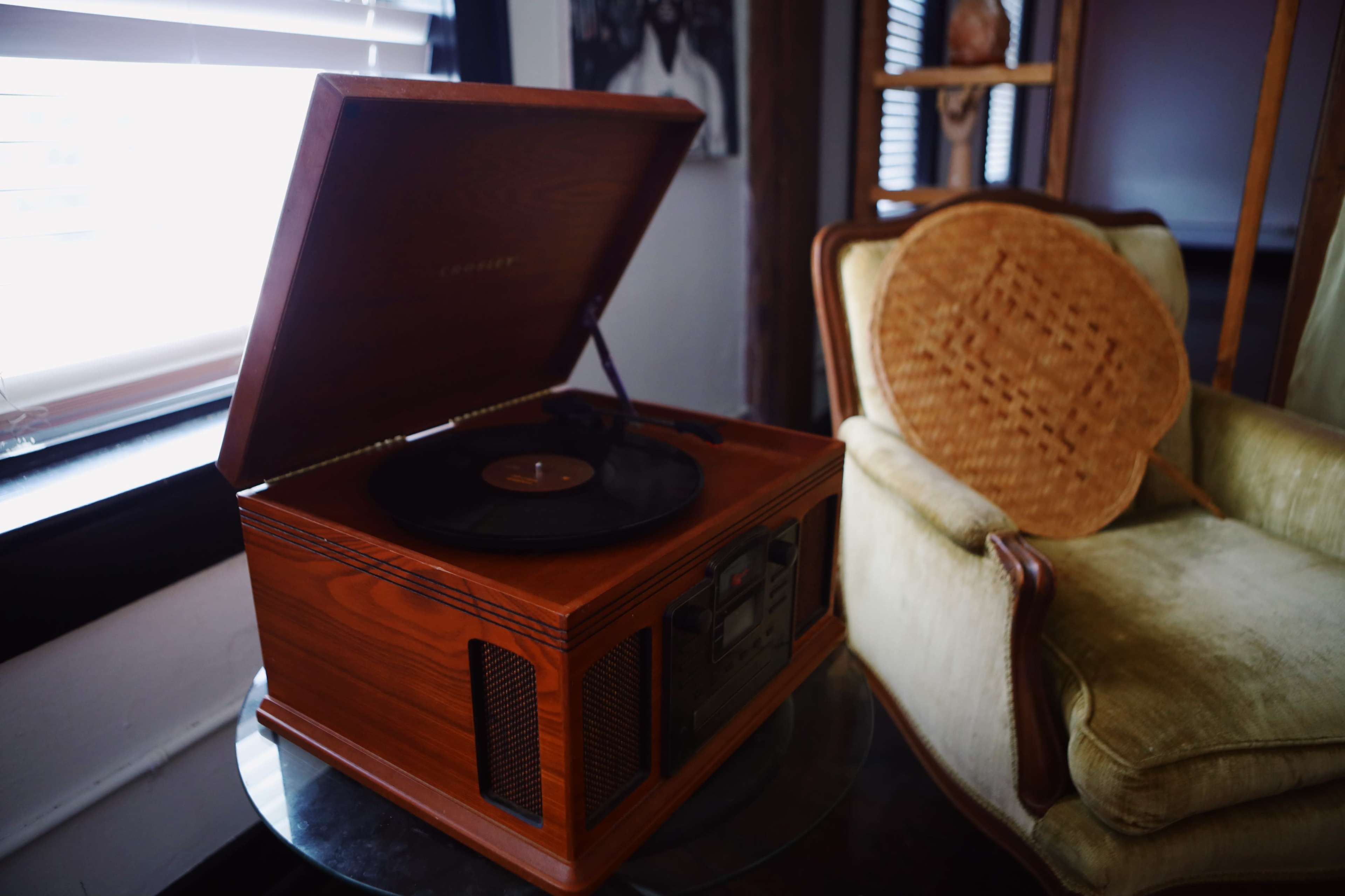 A vintage wooden record player sits on a glass table next to an upholstered chair in a well-lit room.
