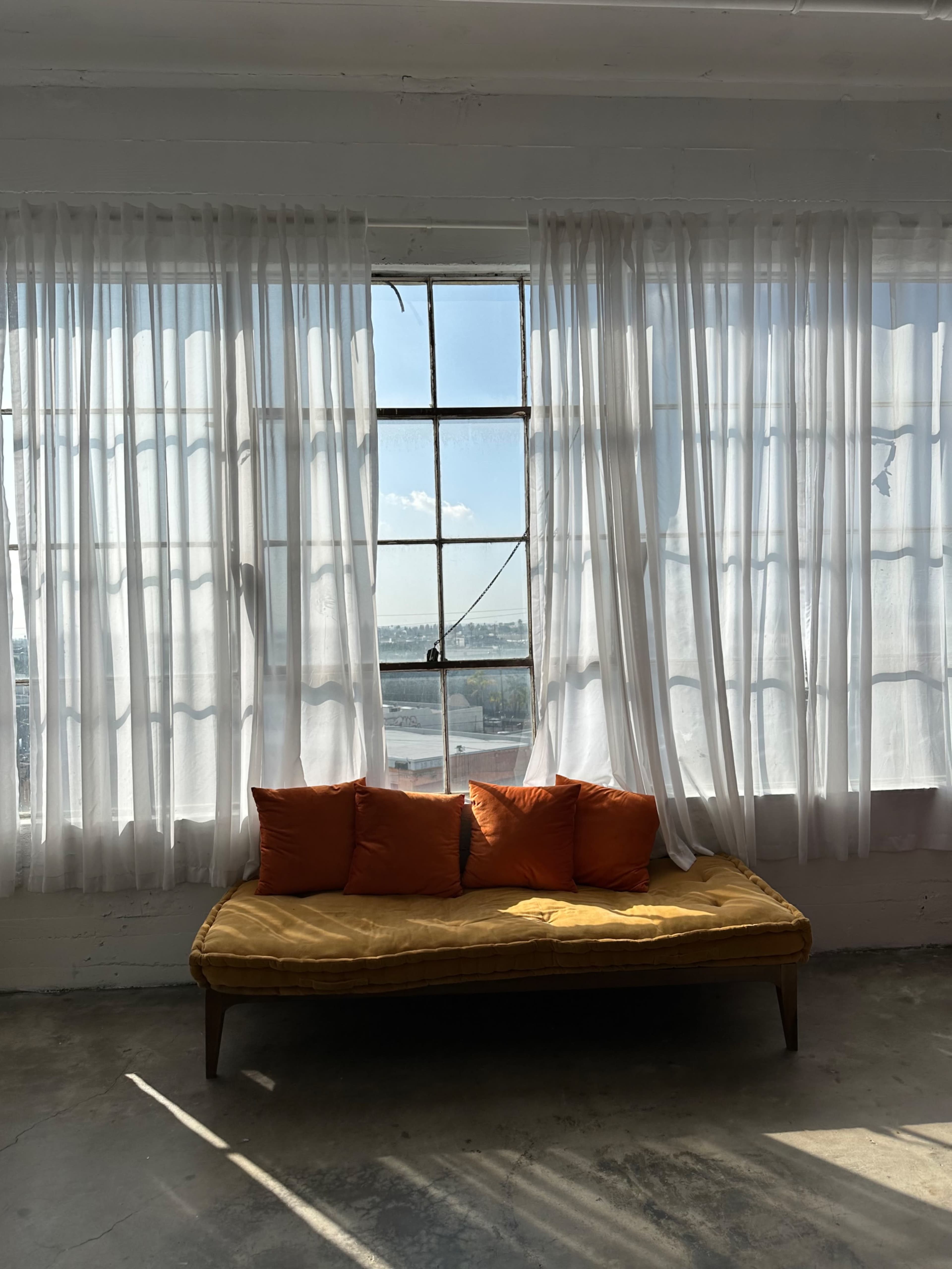 A yellow couch with four orange pillows sits against a large window adorned with sheer white curtains, allowing natural light to fill the space.