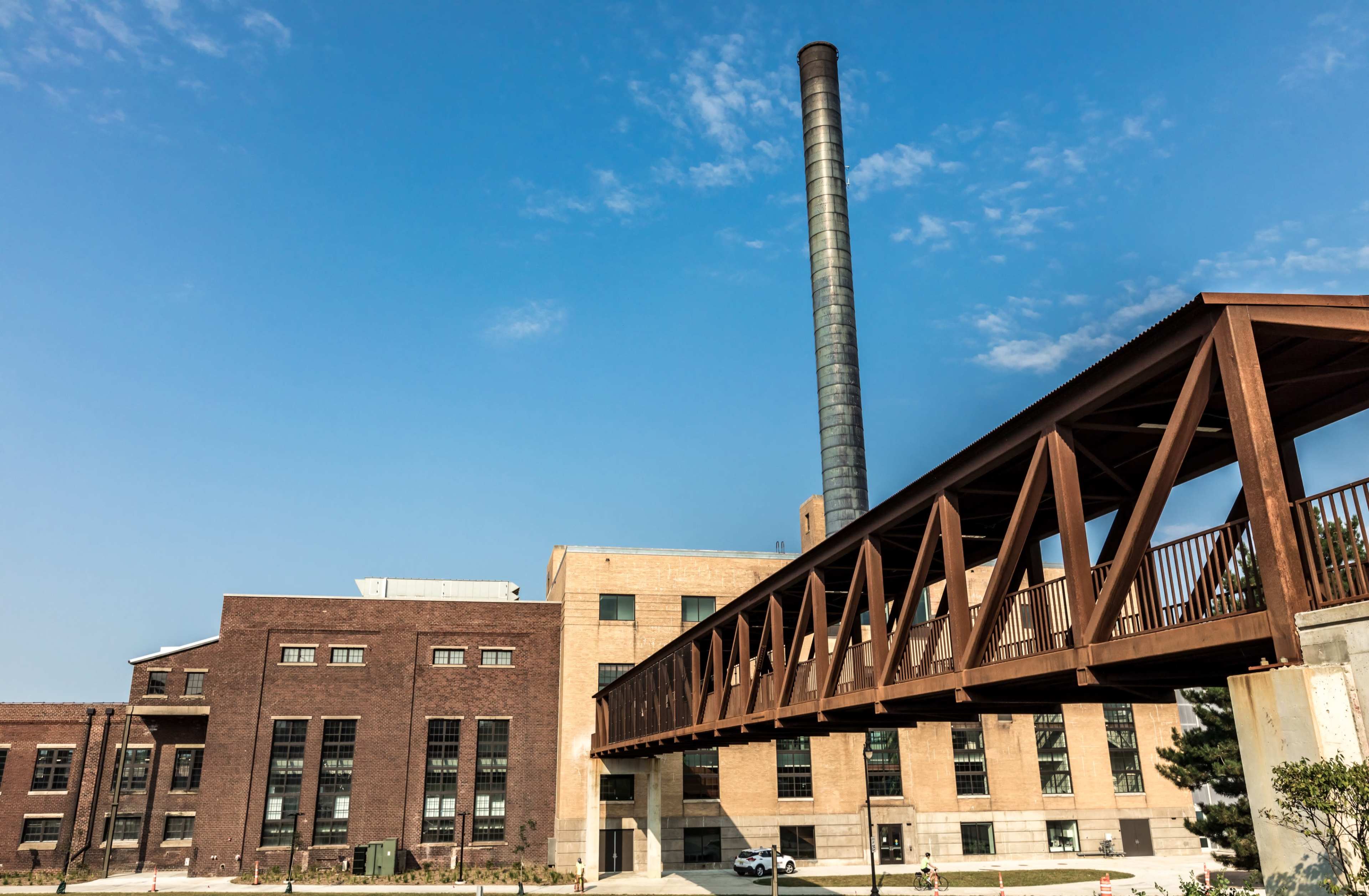 A brick industrial building with a tall smokestack and a brown pedestrian bridge connecting to another structure under a clear blue sky.