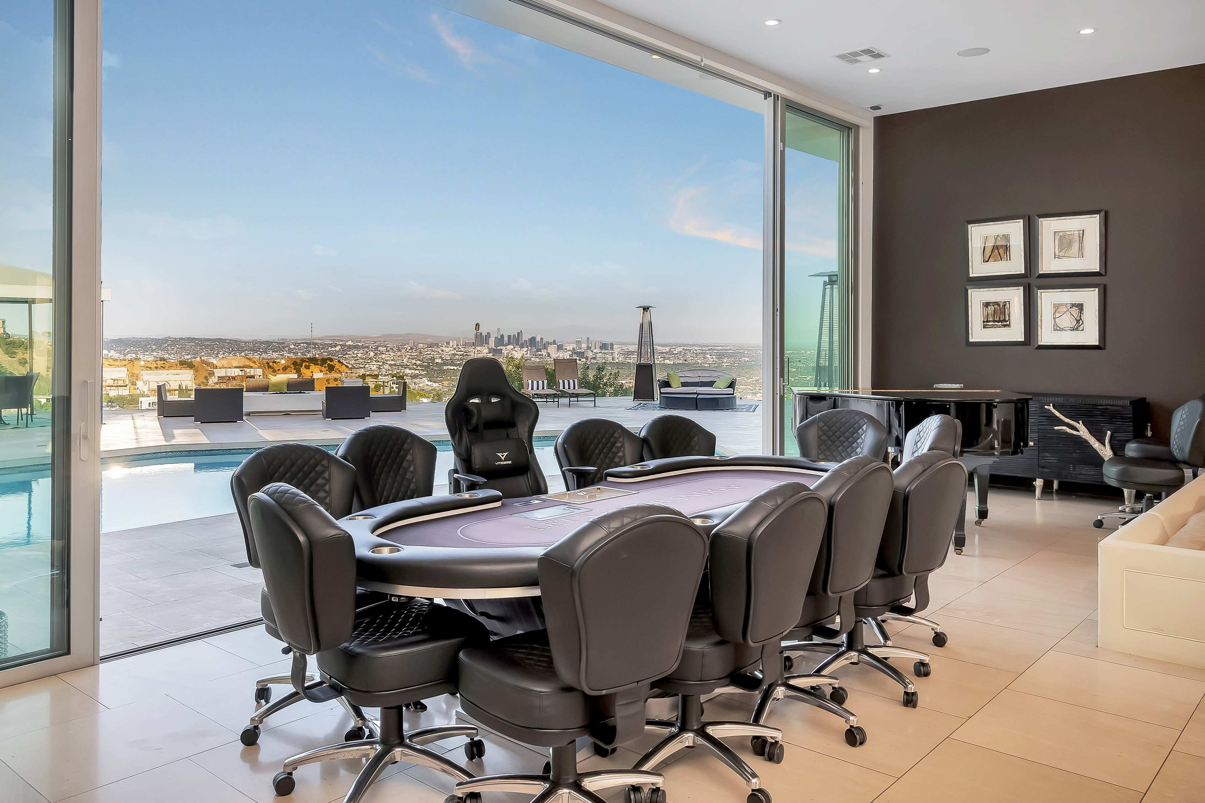 A modern poker room features a large table surrounded by black chairs, with floor-to-ceiling windows offering a view of the city skyline and swimming pool.