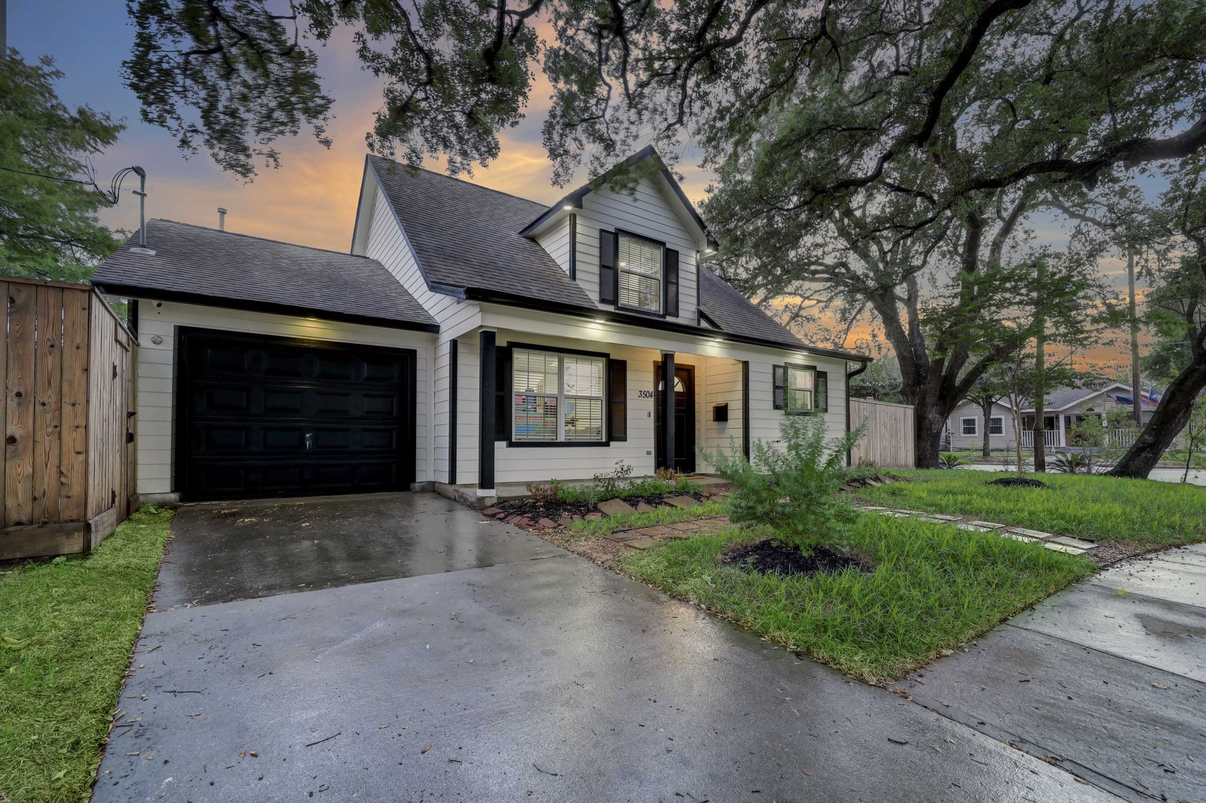 A two-story house with a gable roof, black garage door, and a stone pathway is situated on a wet driveway in a suburban neighborhood during sunset.