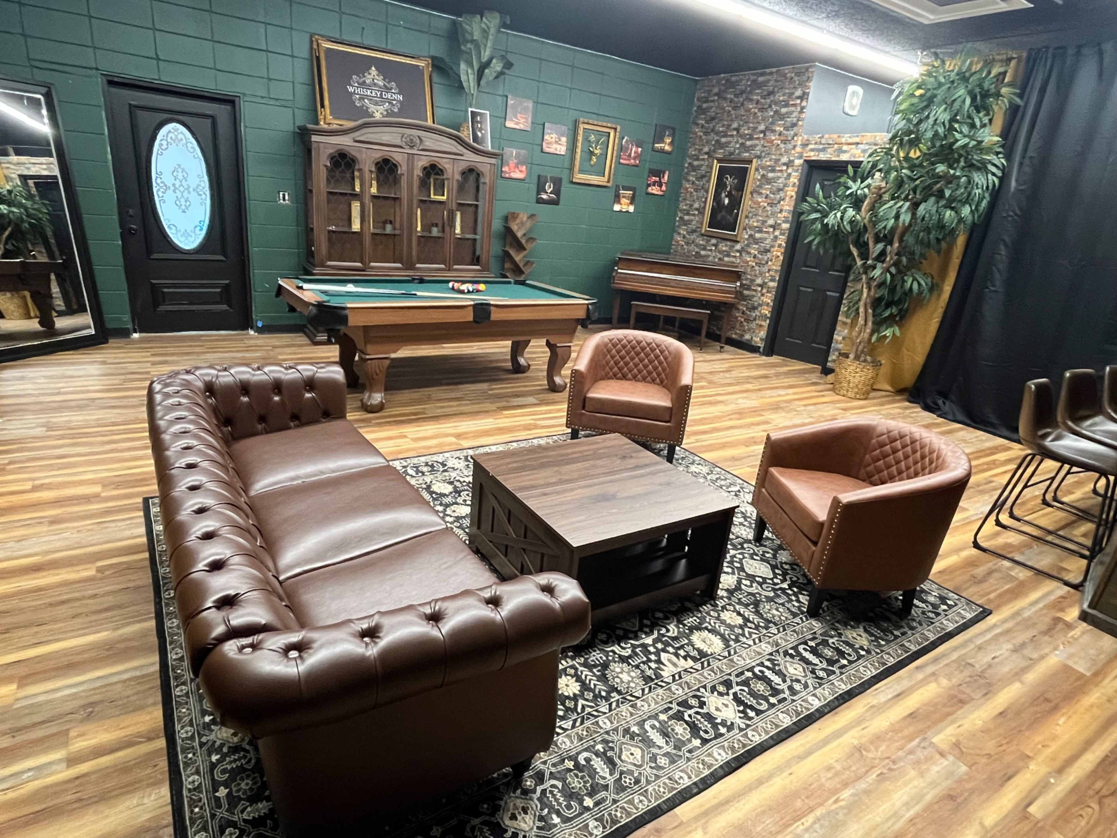 The image shows a game room featuring a billiards table, a brown leather couch and chairs, a decorative wooden cabinet, and various framed pictures on the walls.