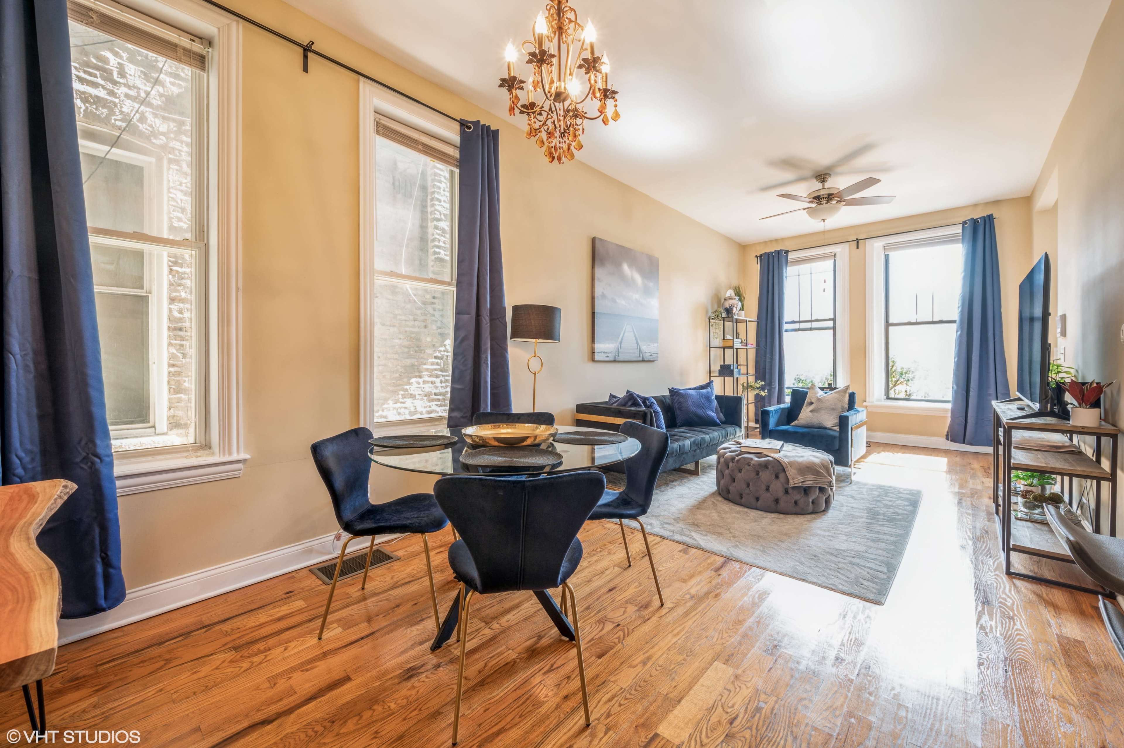 A modern living room featuring a glass dining table, dark blue curtains, a comfortable seating area, and large windows allowing natural light to fill the space.