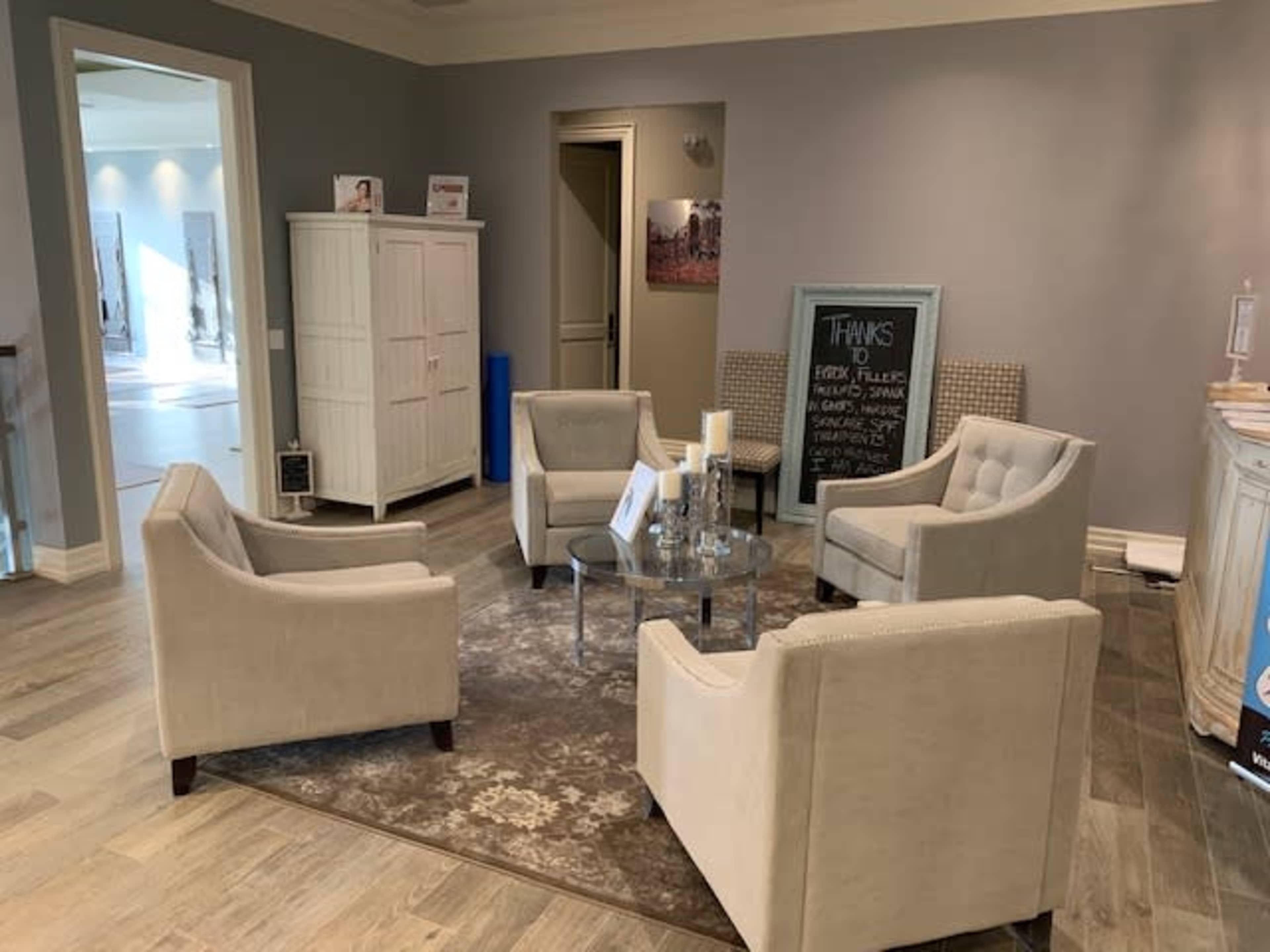 The image shows a small waiting area with four upholstered chairs arranged around a glass coffee table on a patterned rug, against a light gray wall.