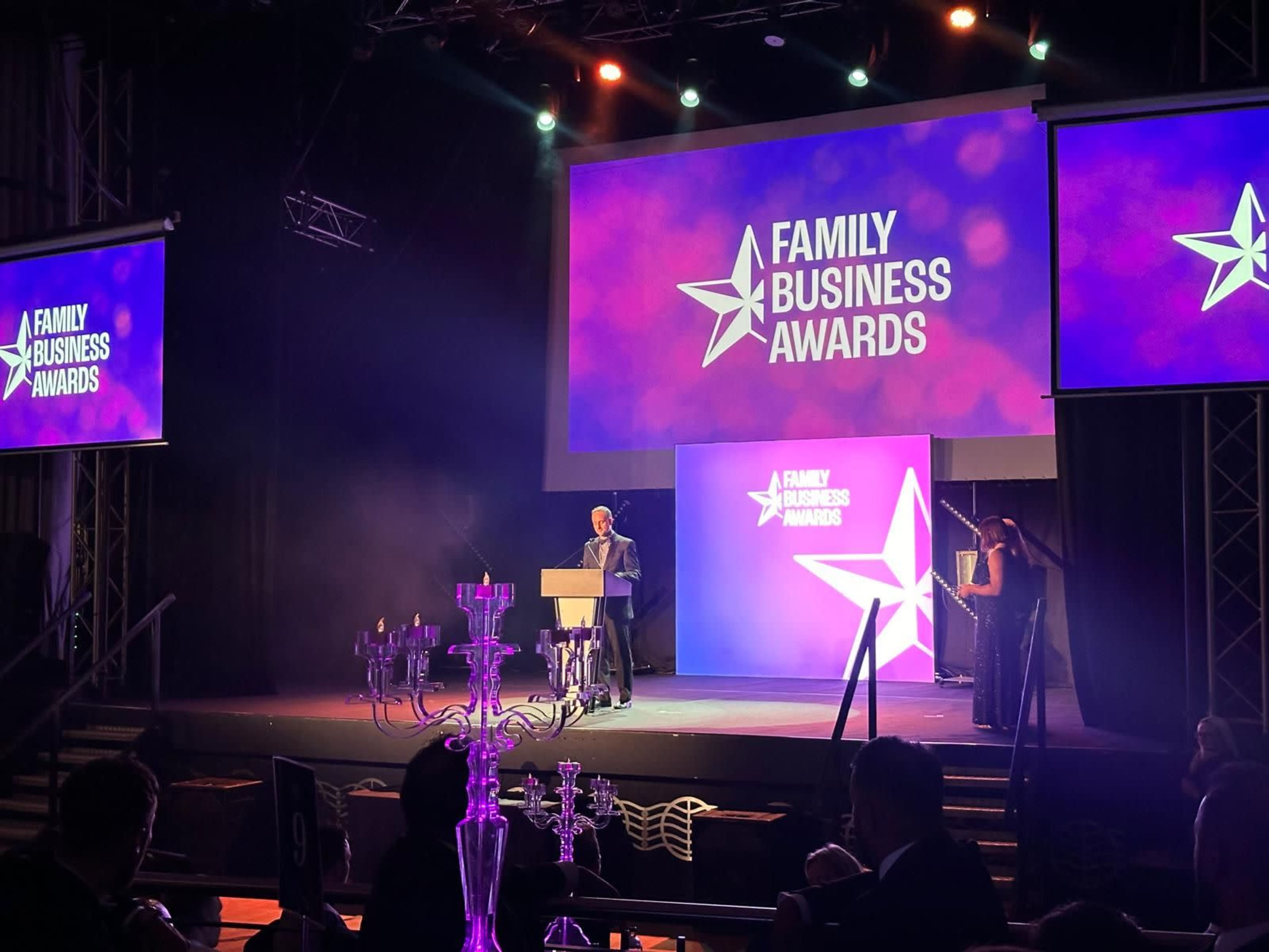 A speaker is addressing the audience at the Family Business Awards event, with colorful screens displaying the event's logo in a dimly lit venue.