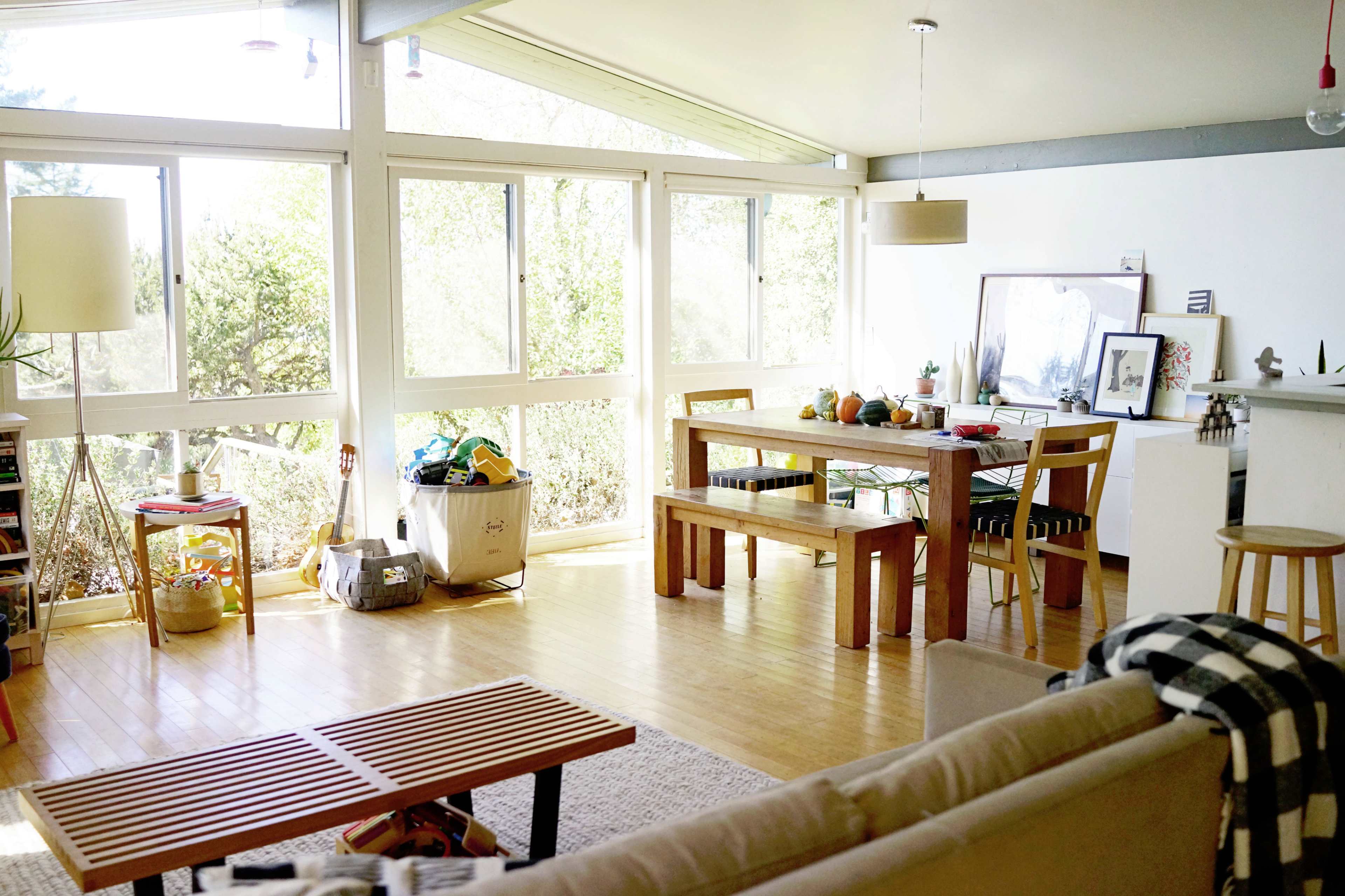 A bright, open living area features large windows, a wooden dining table, a sofa, and decorative items throughout.