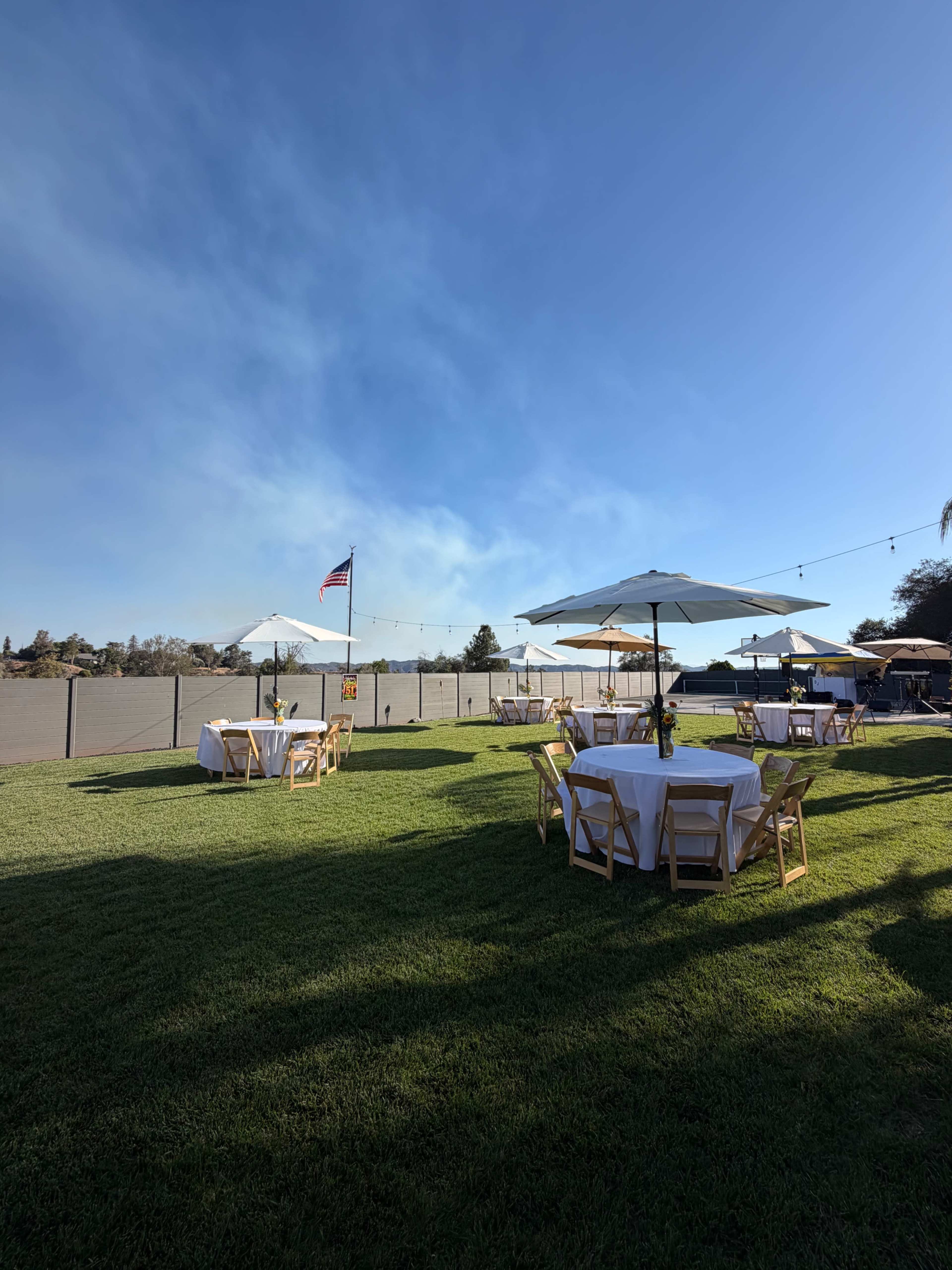 The image shows a grassy outdoor area with wooden tables set for dining under large umbrellas, against a clear blue sky and a distant flag.