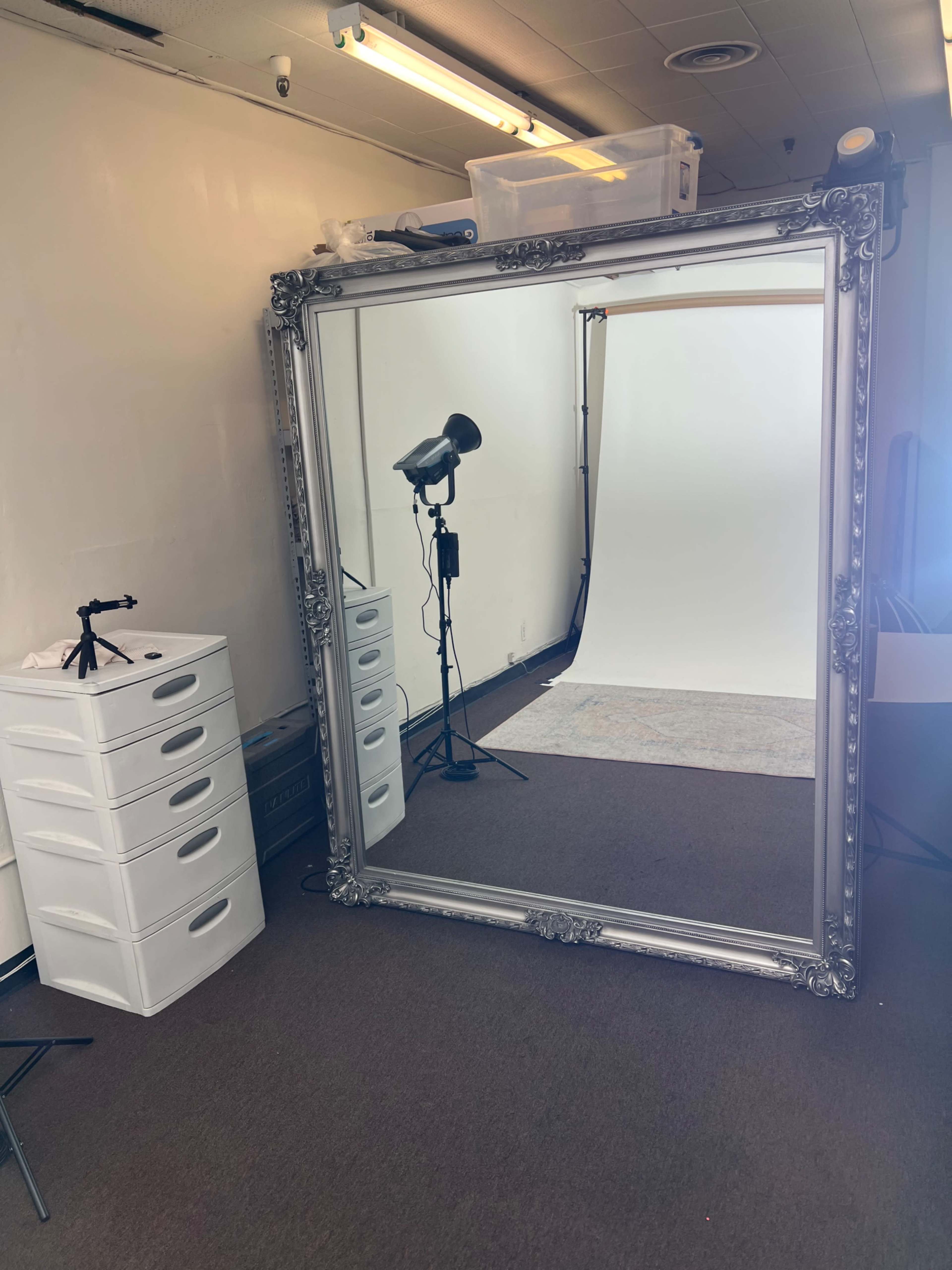 The image shows a large ornate silver mirror leaning against a wall in a photo studio, with a white backdrop and a light setup visible in the background.