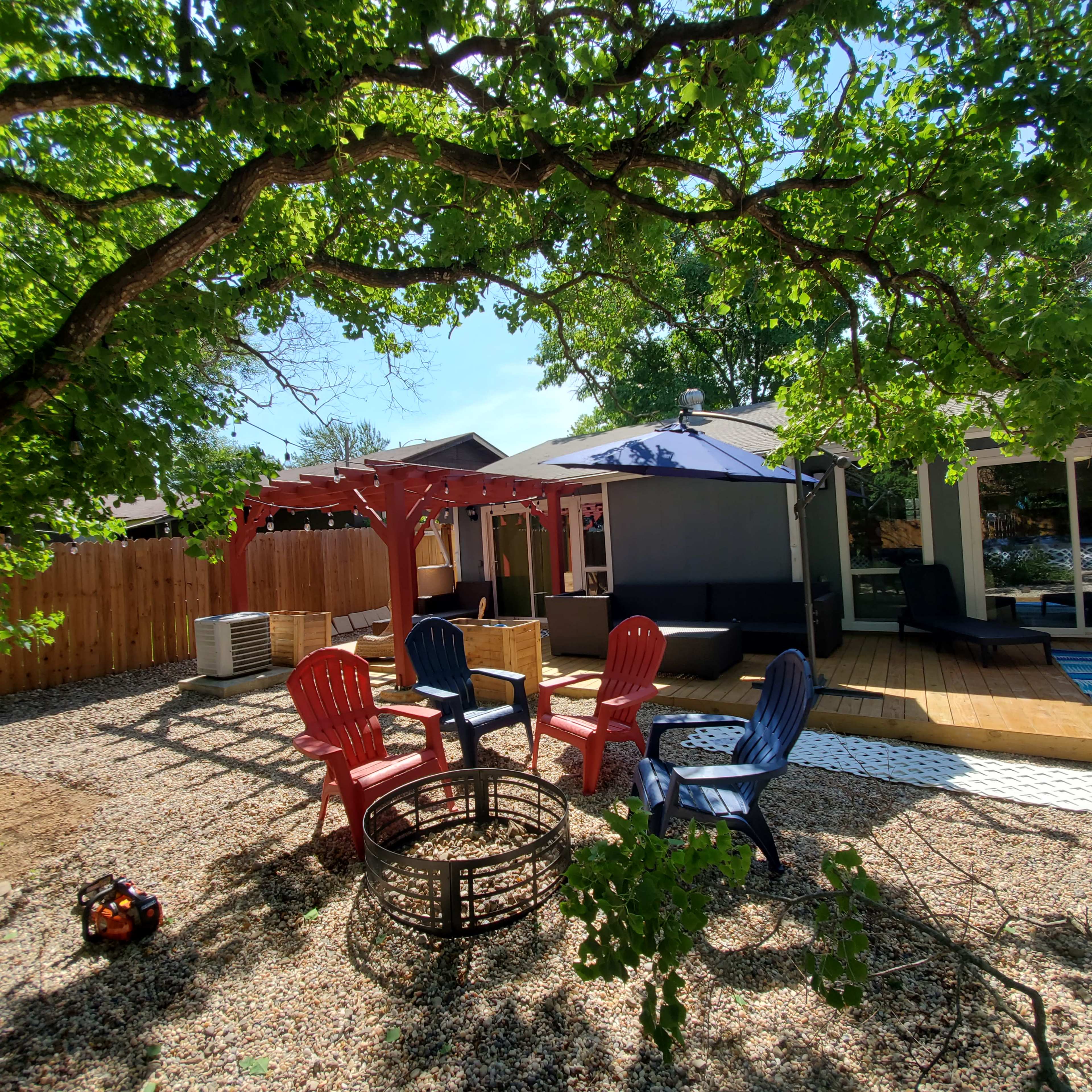 A backyard area with a fire pit surrounded by colorful chairs, a wooden deck, and a shaded seating space under a pergola.