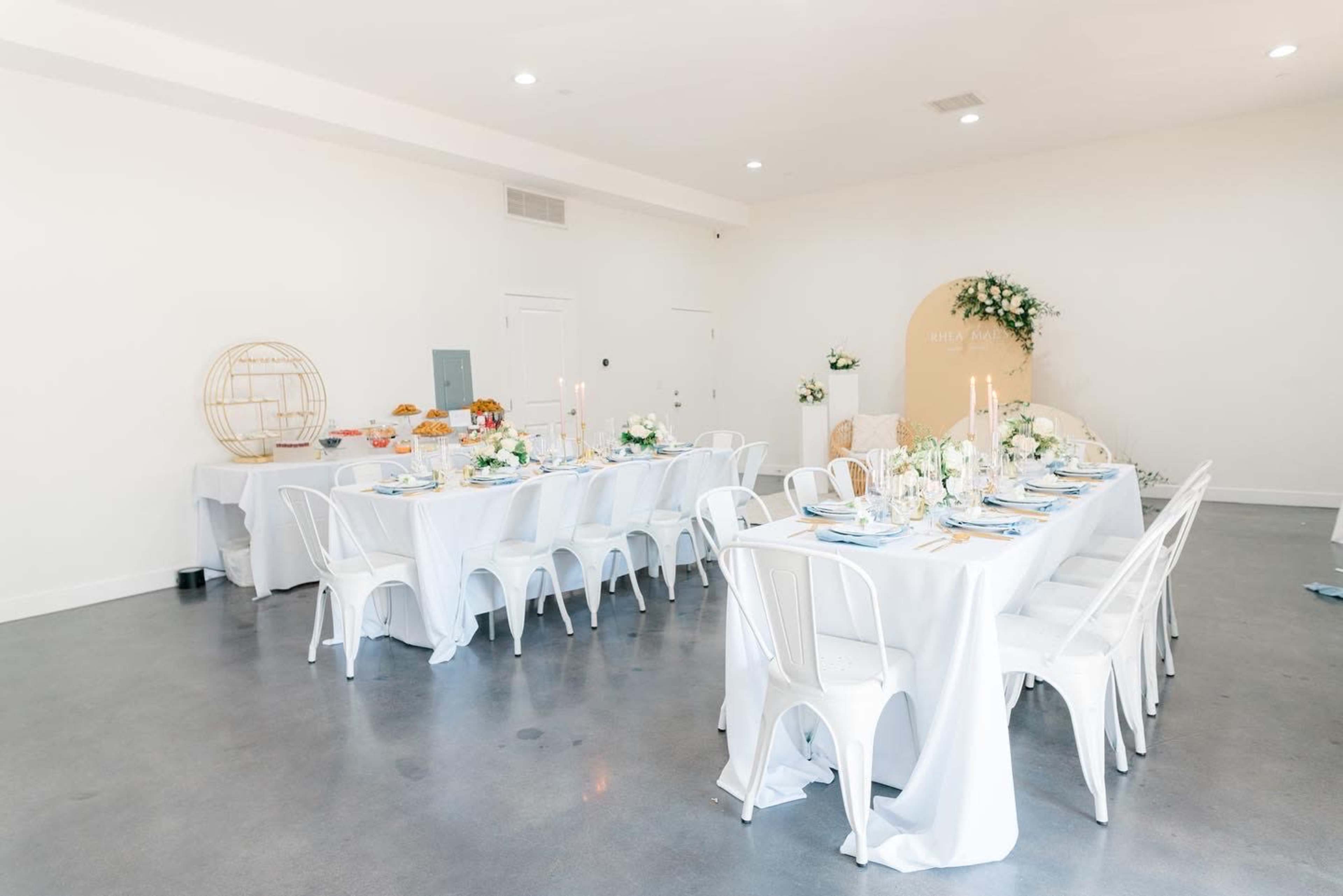 A spacious, elegantly arranged dining area features white tablecloths, multiple tables set with dishware, and a decorative backdrop adorned with flowers.