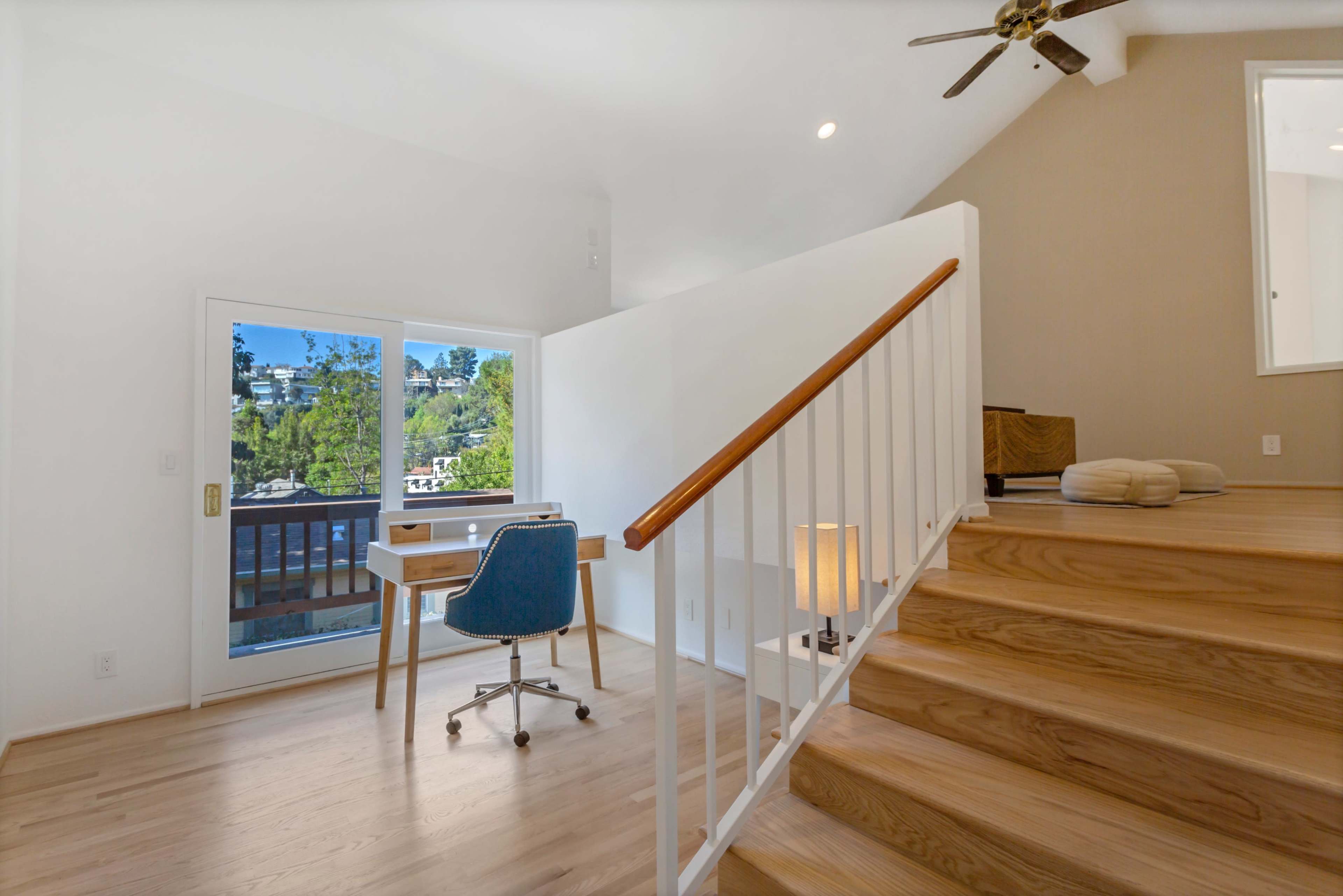 A bright, airy interior features a wooden staircase leading to a small desk with a blue chair beside a window overlooking a green landscape.