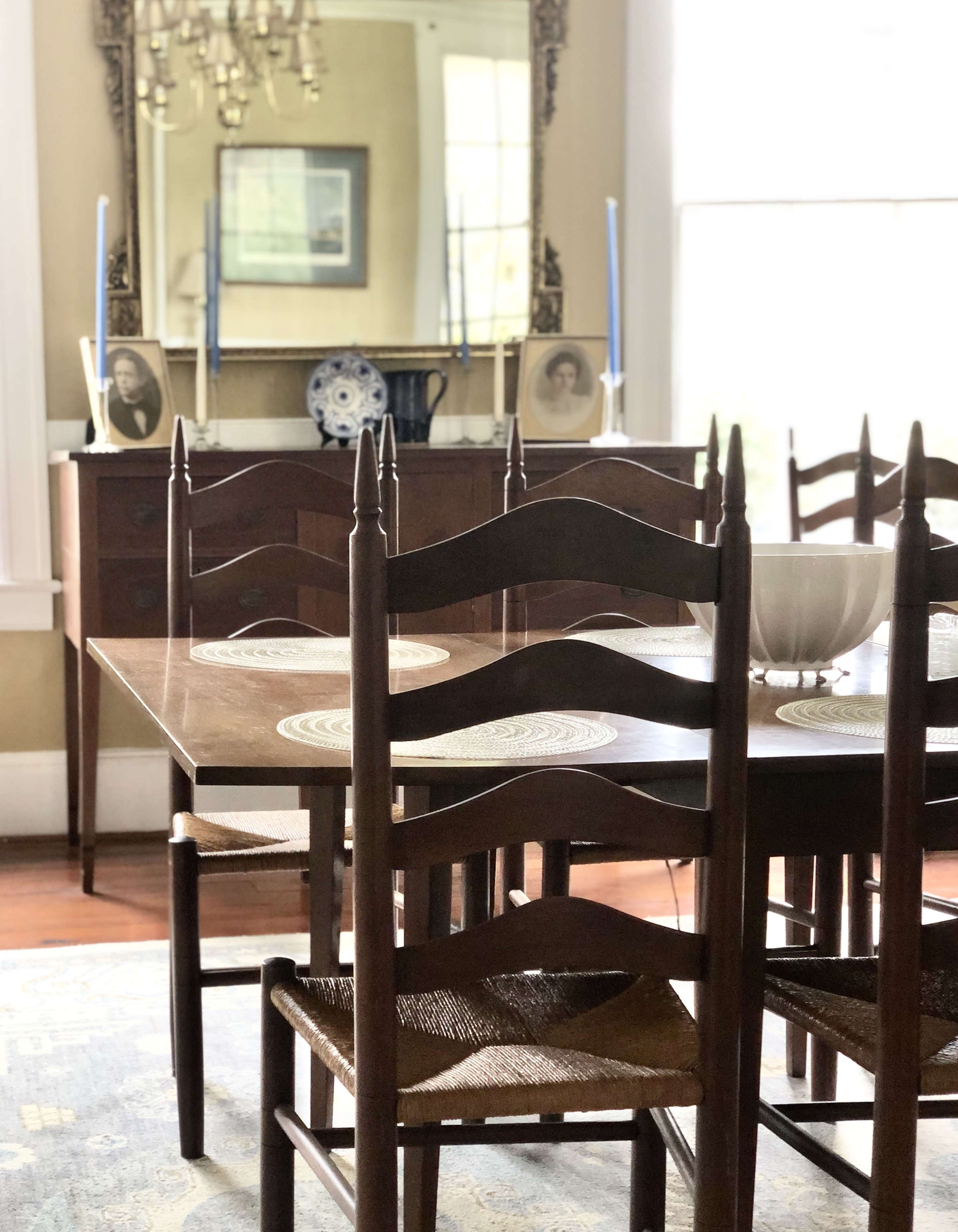 The image depicts a dimly lit dining room featuring a wooden table surrounded by chairs, with a mirror and decorative items on a sideboard in the background.