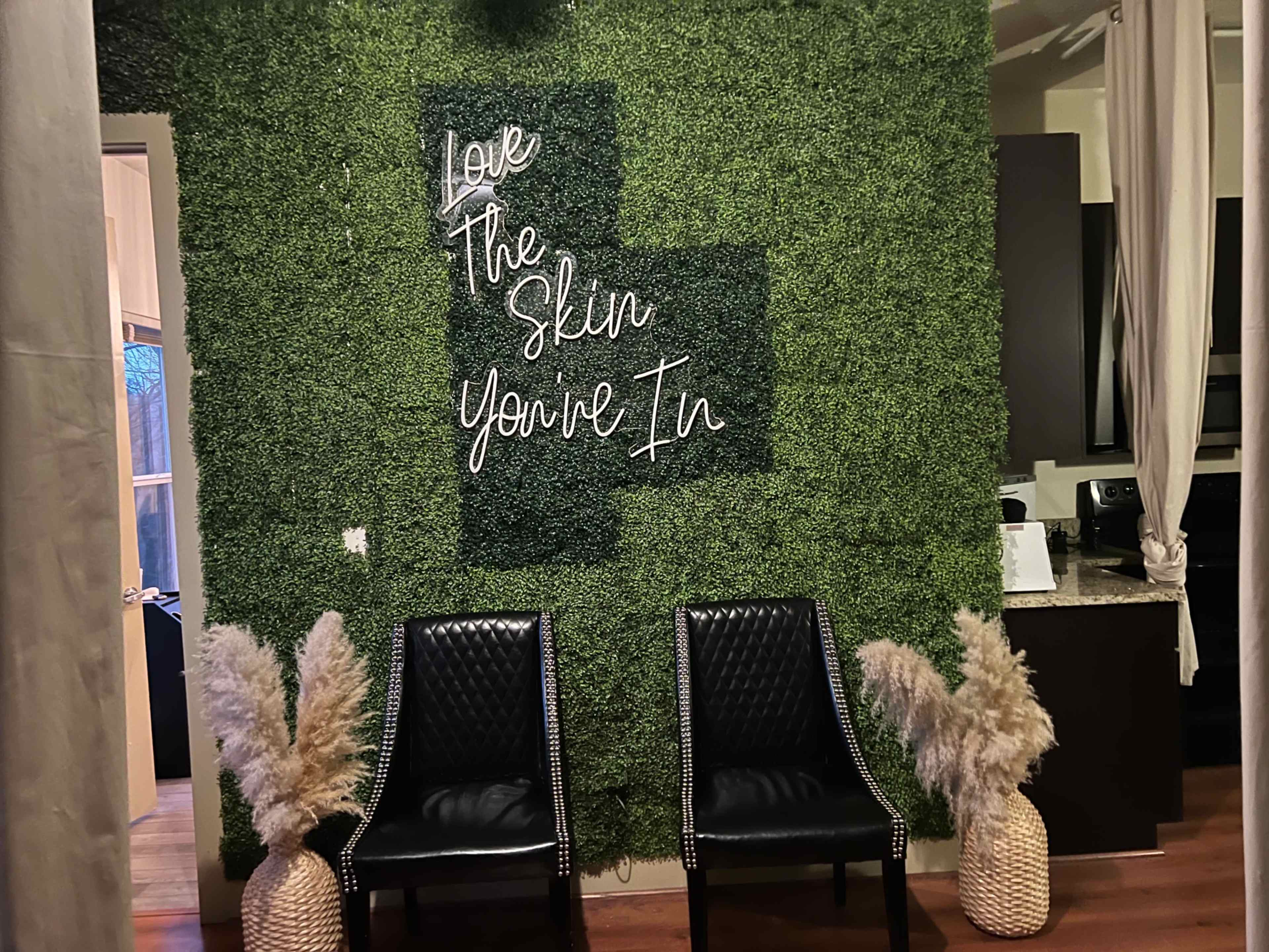 A green wall with a motivational quote, "Love The Skin You're In," and two black chairs flanking a decorative arrangement of dried pampas grass.