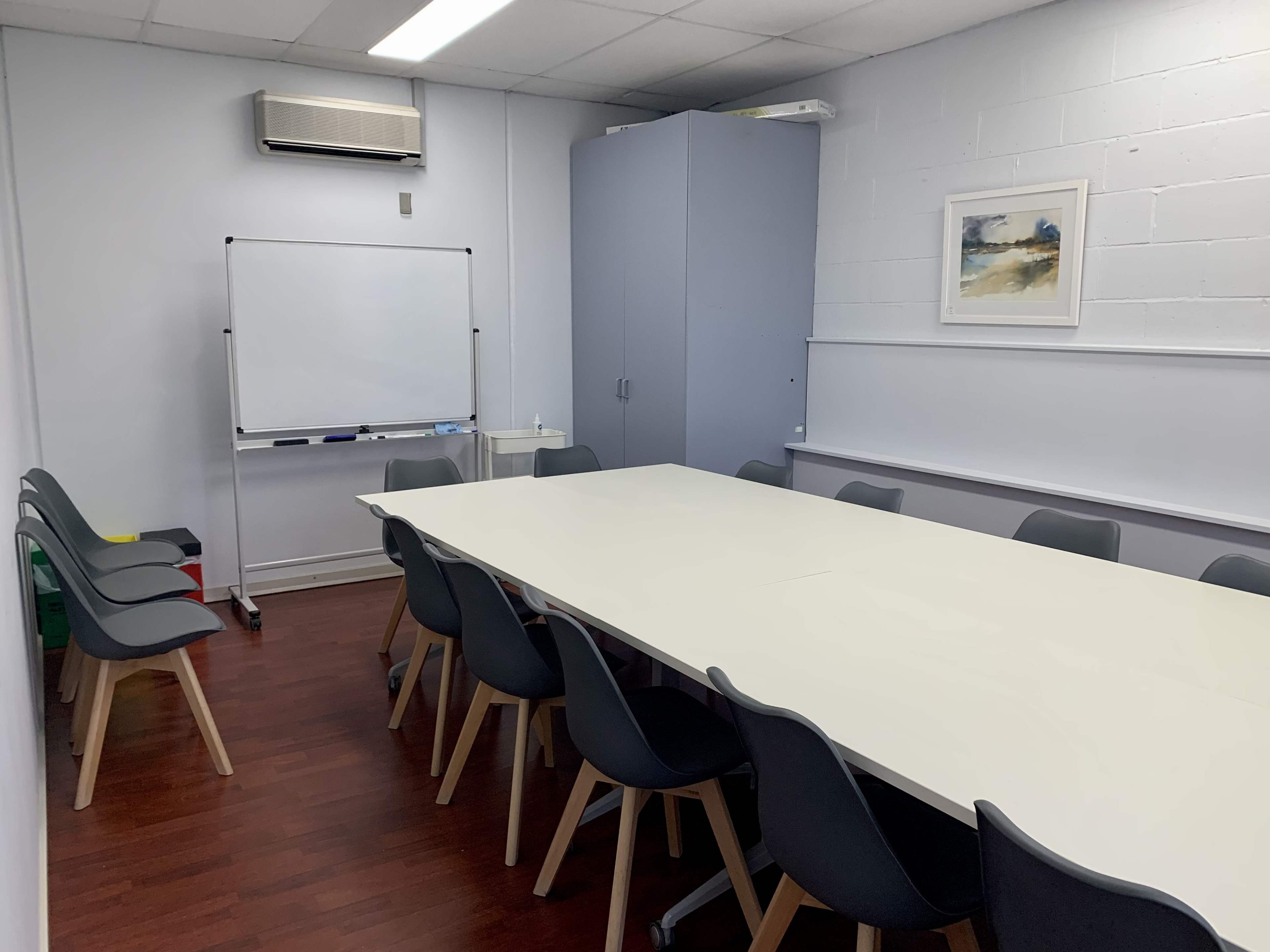 The image shows a meeting room with a large white table surrounded by gray chairs, a whiteboard, and a cabinet.