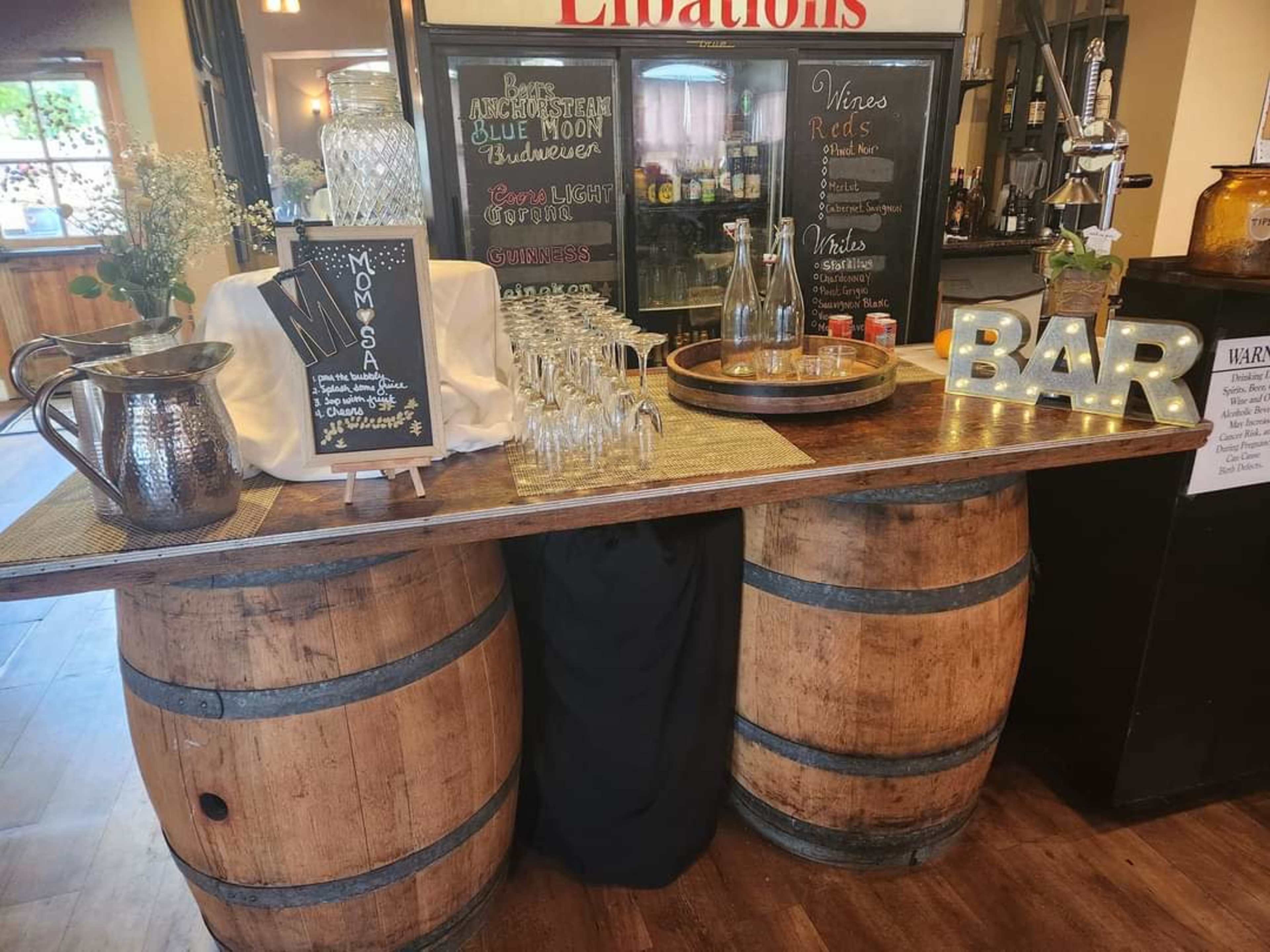 A bar area made from two stacked wooden barrels, featuring a menu board for mimosas, a selection of glasses, and decorative lighting.