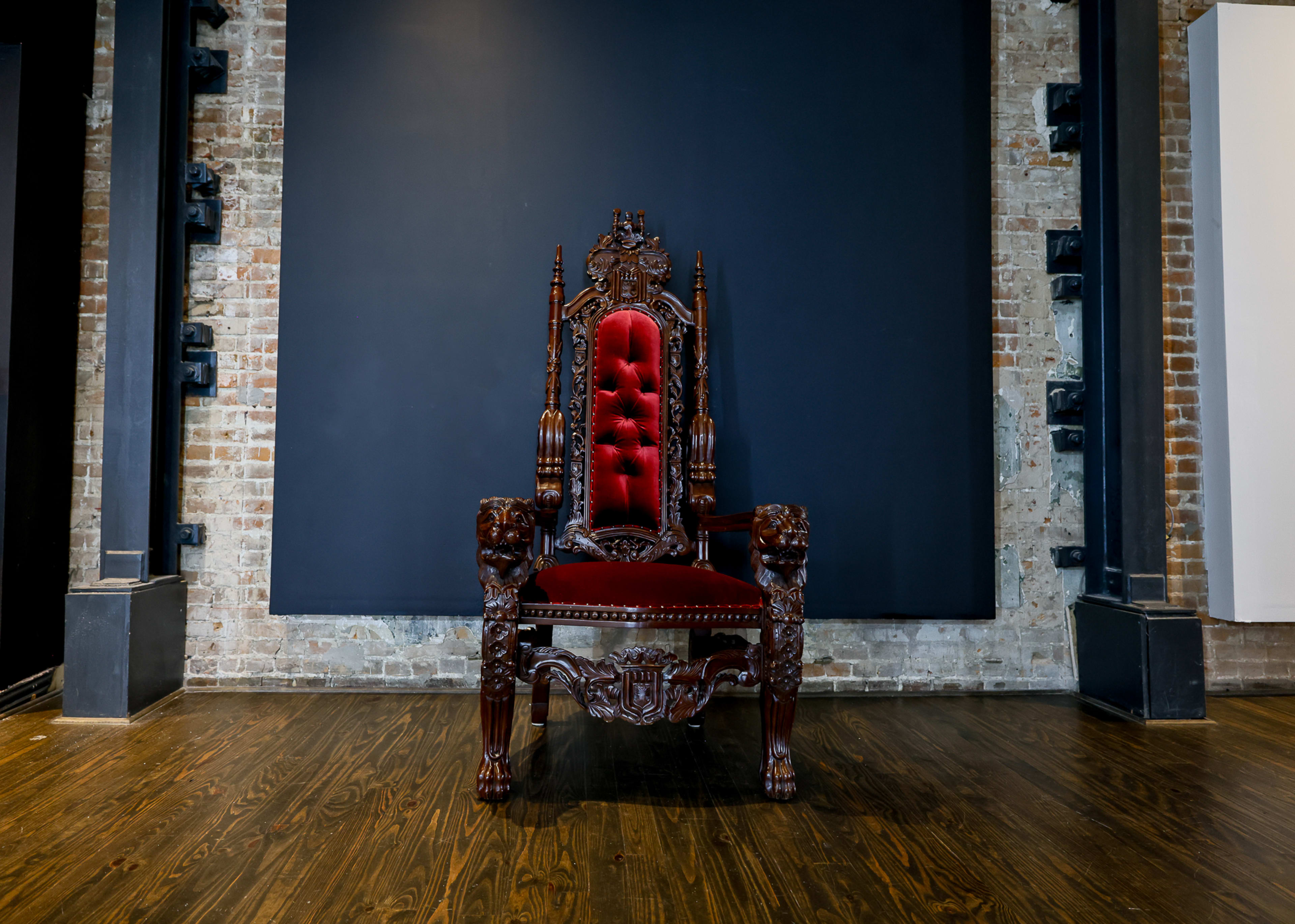 A dark wood throne with red upholstery is centered in front of a large black wall in an industrial-style room.