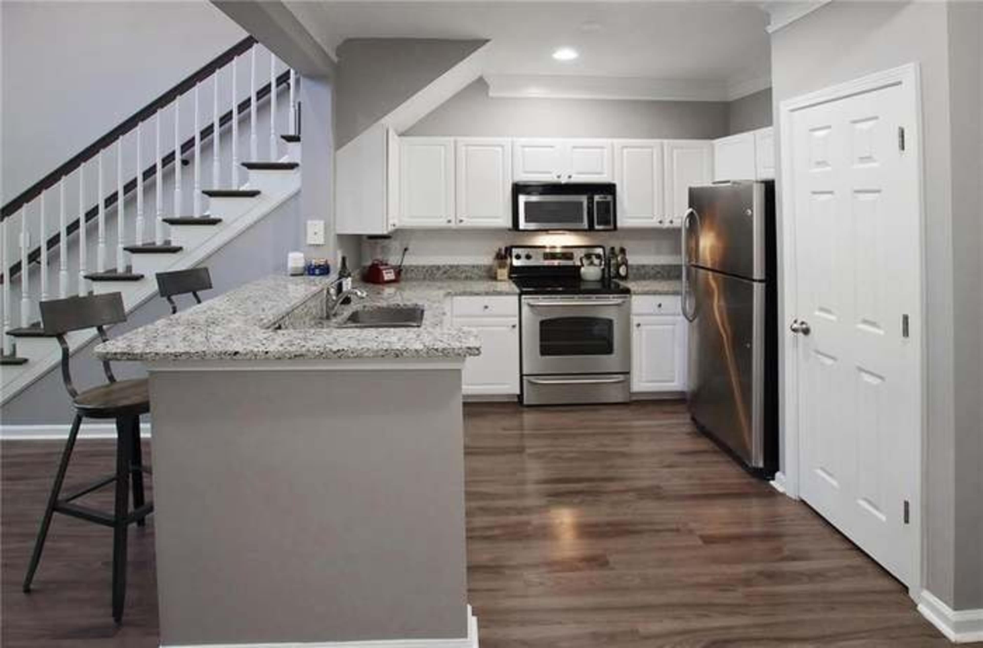 A modern kitchen with white cabinets, stainless steel appliances, and a granite countertop, adjacent to a staircase with wooden steps.