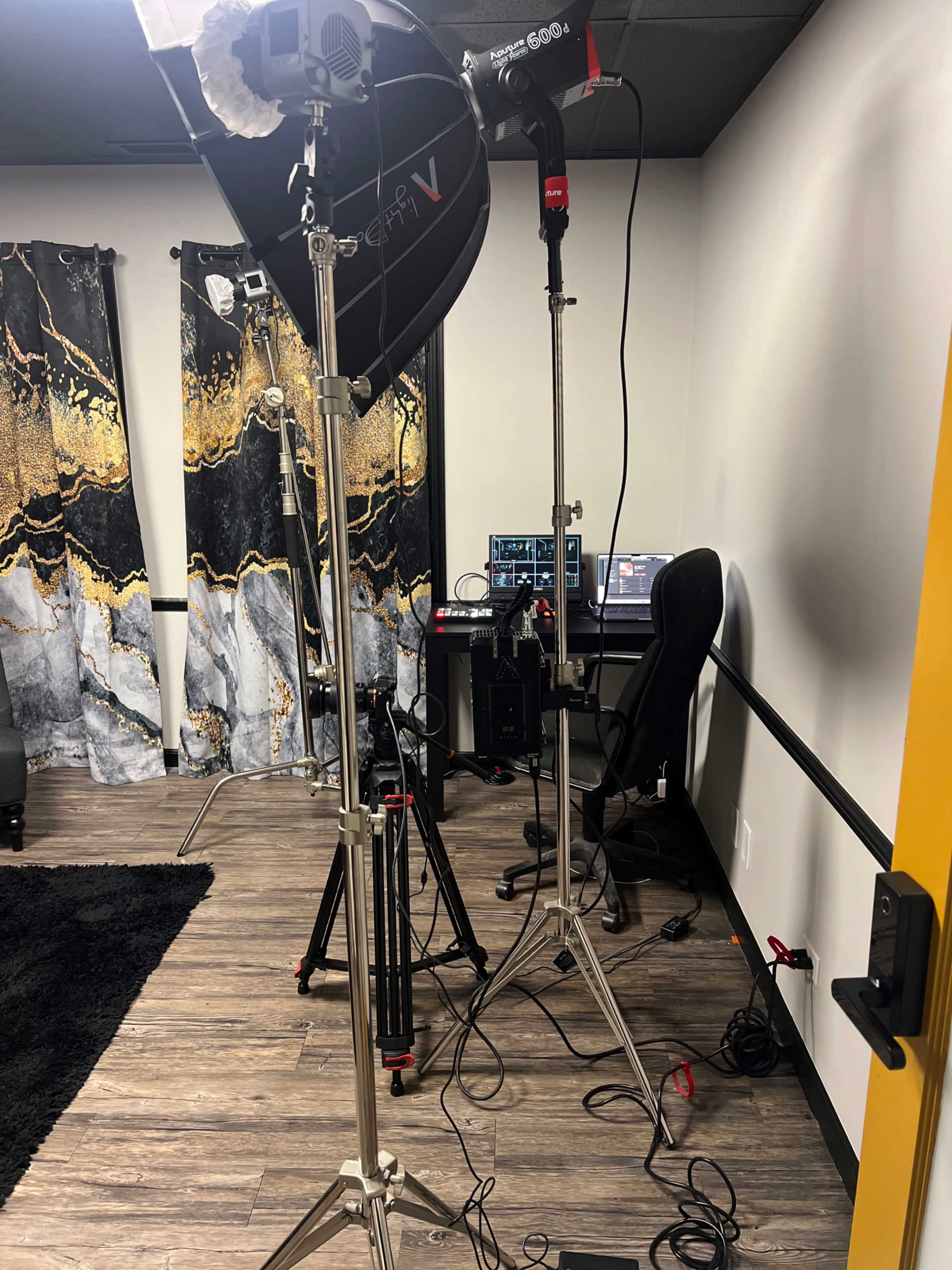 The image shows a photography studio setup with lighting equipment, a camera tripod, and a desk with a computer in the background.